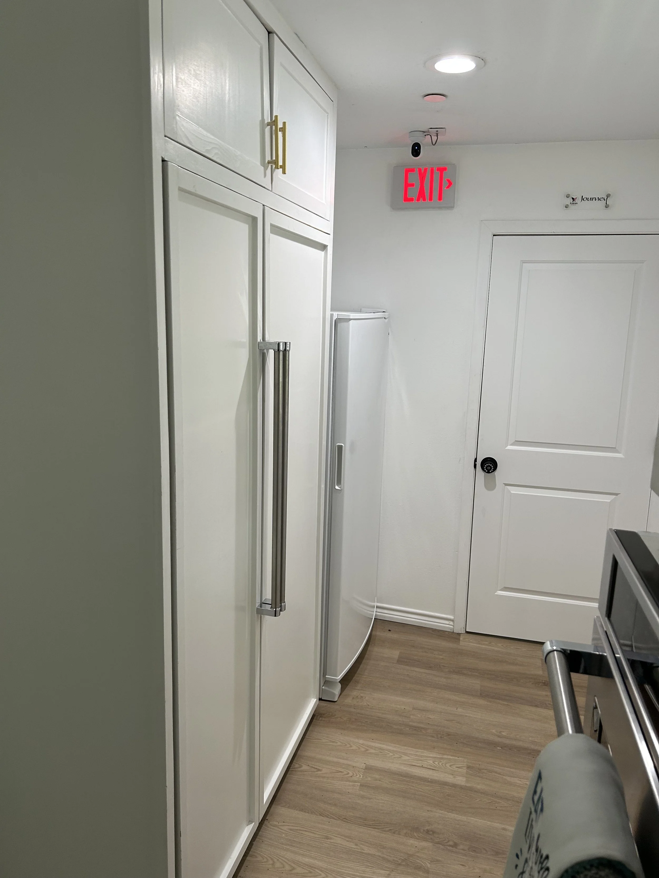 A white refrigerator with a handle is located next to a white door in a room with wood-like flooring. An exit sign is mounted on the wall above the door, and a security camera is also mounted on the ceiling nearby.