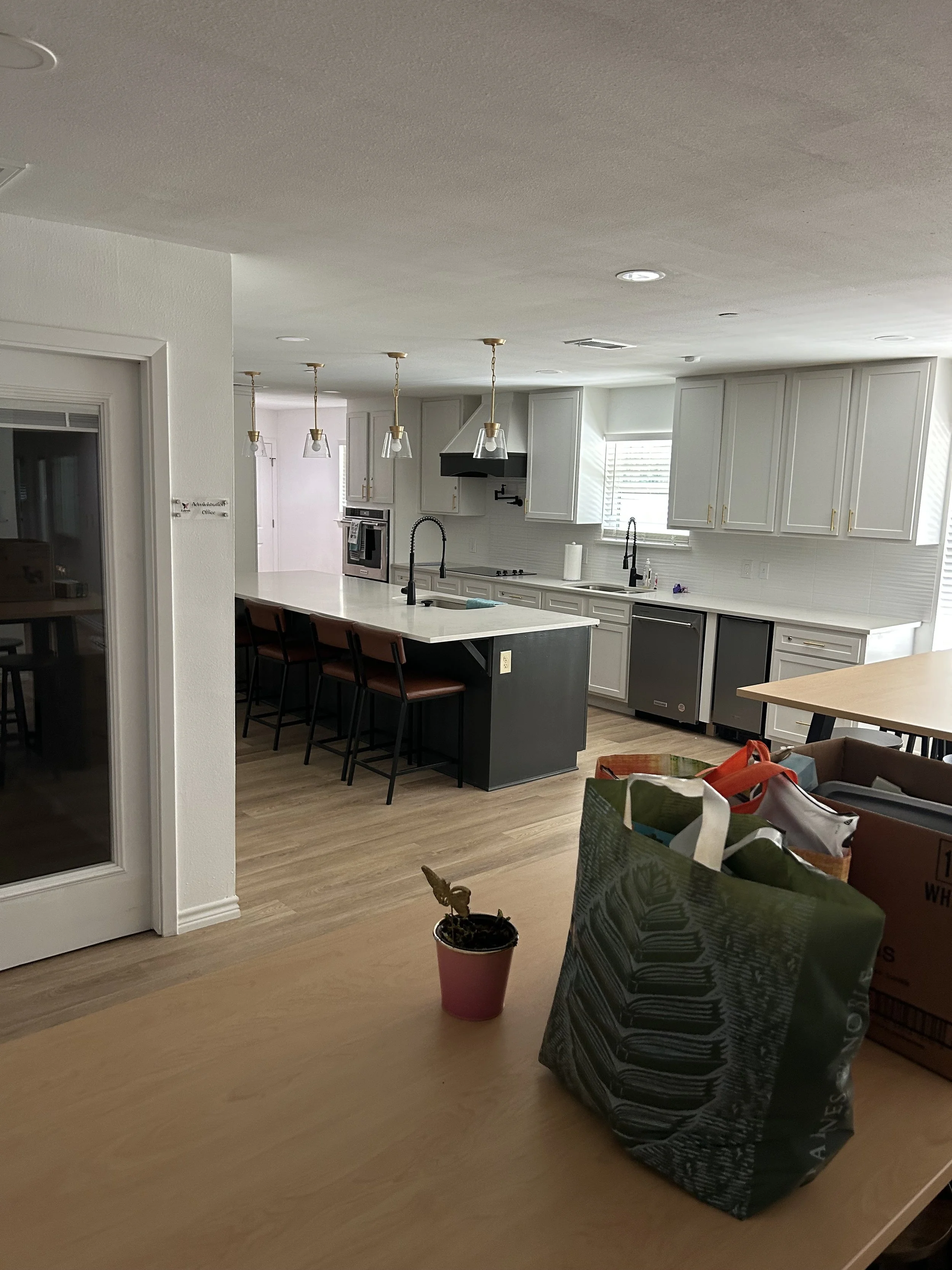 Kitchen with white cabinets, island with bar stools, black sink, black faucet, black range hood, white backsplash, and three hanging pendant lights. Some items and bags are on the table in the foreground.