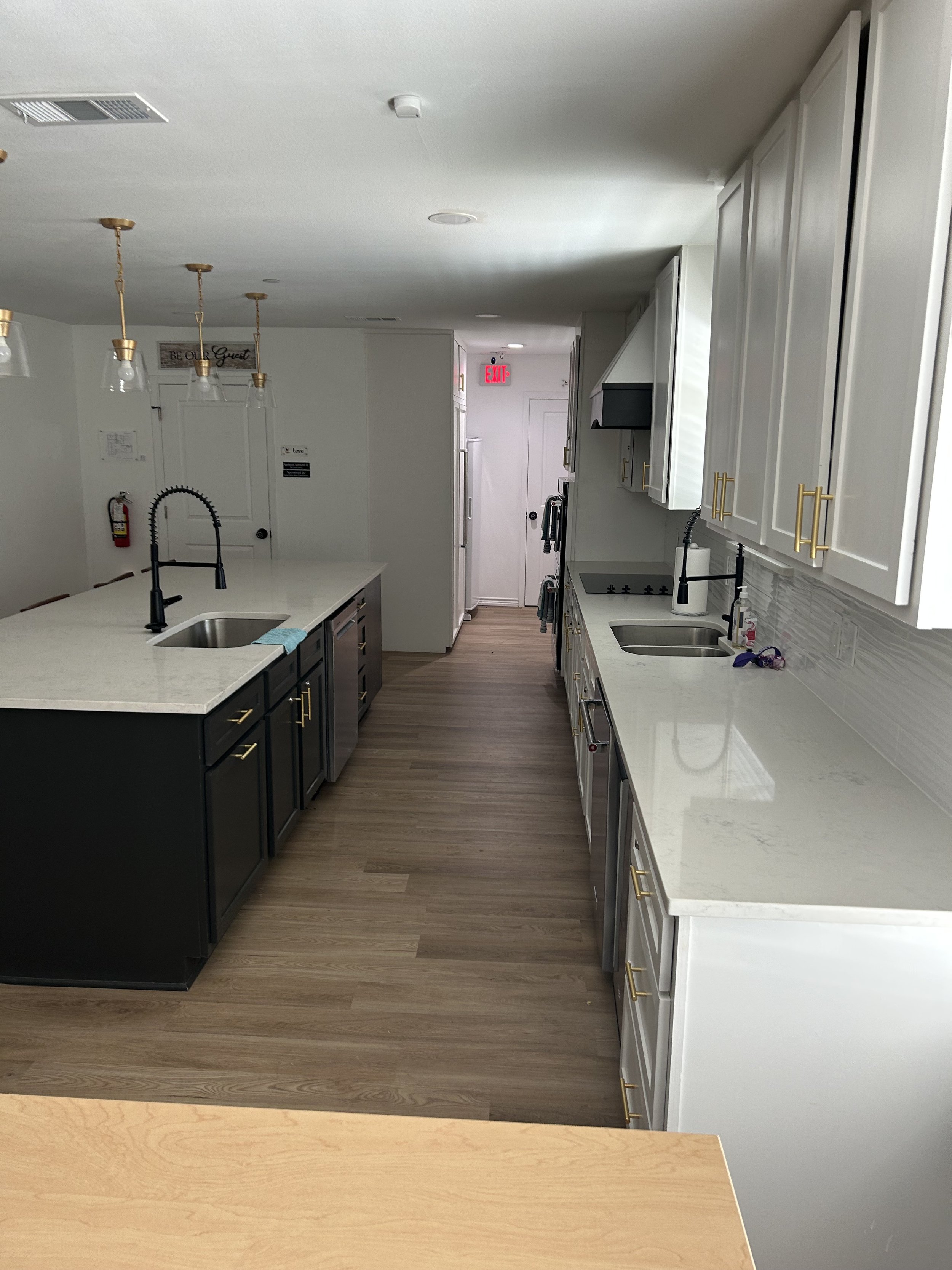 A modern kitchen with white cabinets and black lower cabinets, a white countertop, and stainless steel sinks. There are black and gold fixtures, a white backsplash, and a wood floor. The background includes a doorway, a white refrigerator, and an exi