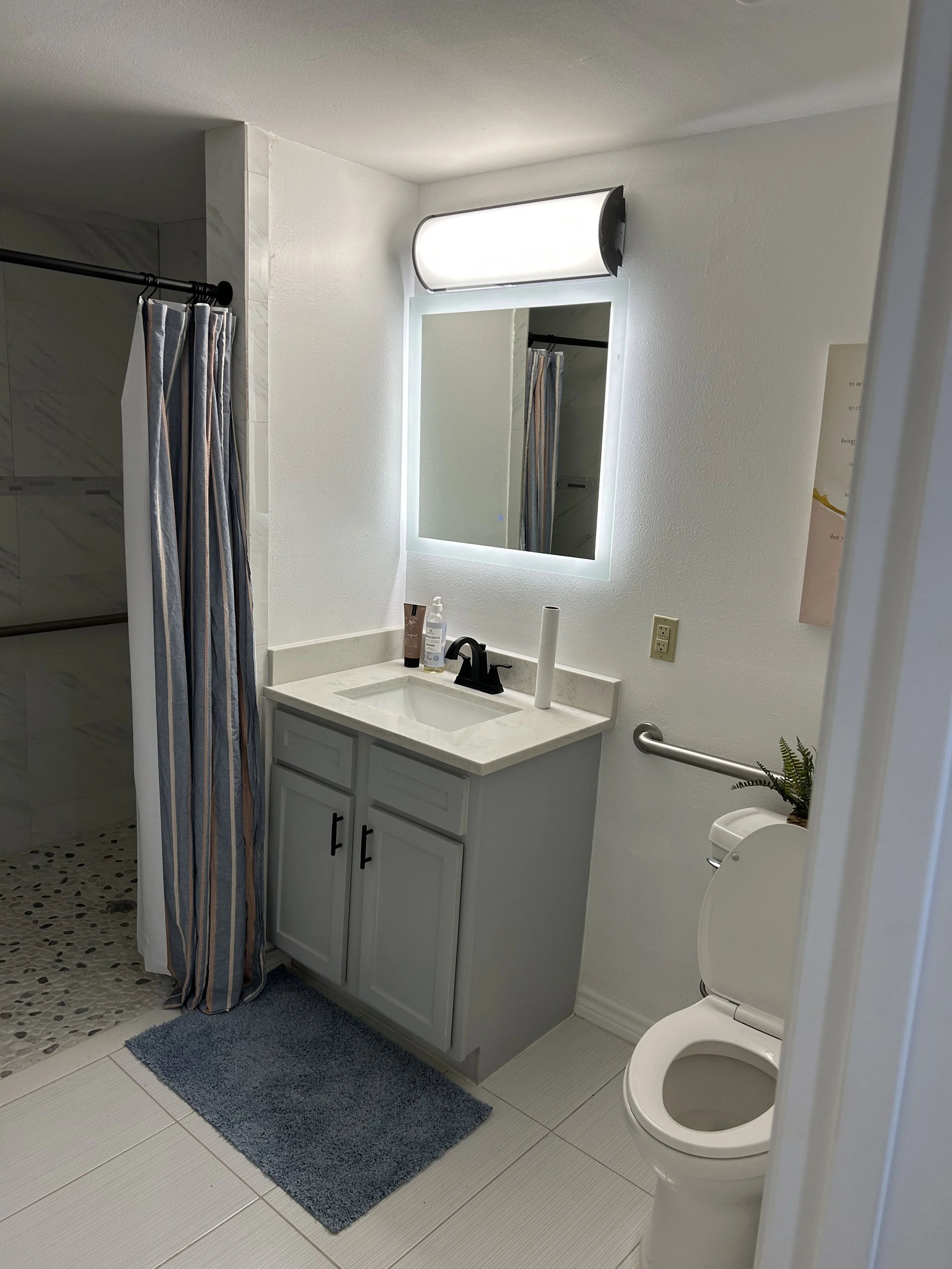 Bathroom with a mirror, lit mirror light, sink, vanity, toiletries, and a toilet, with a shower area visible on the left with a shower curtain.