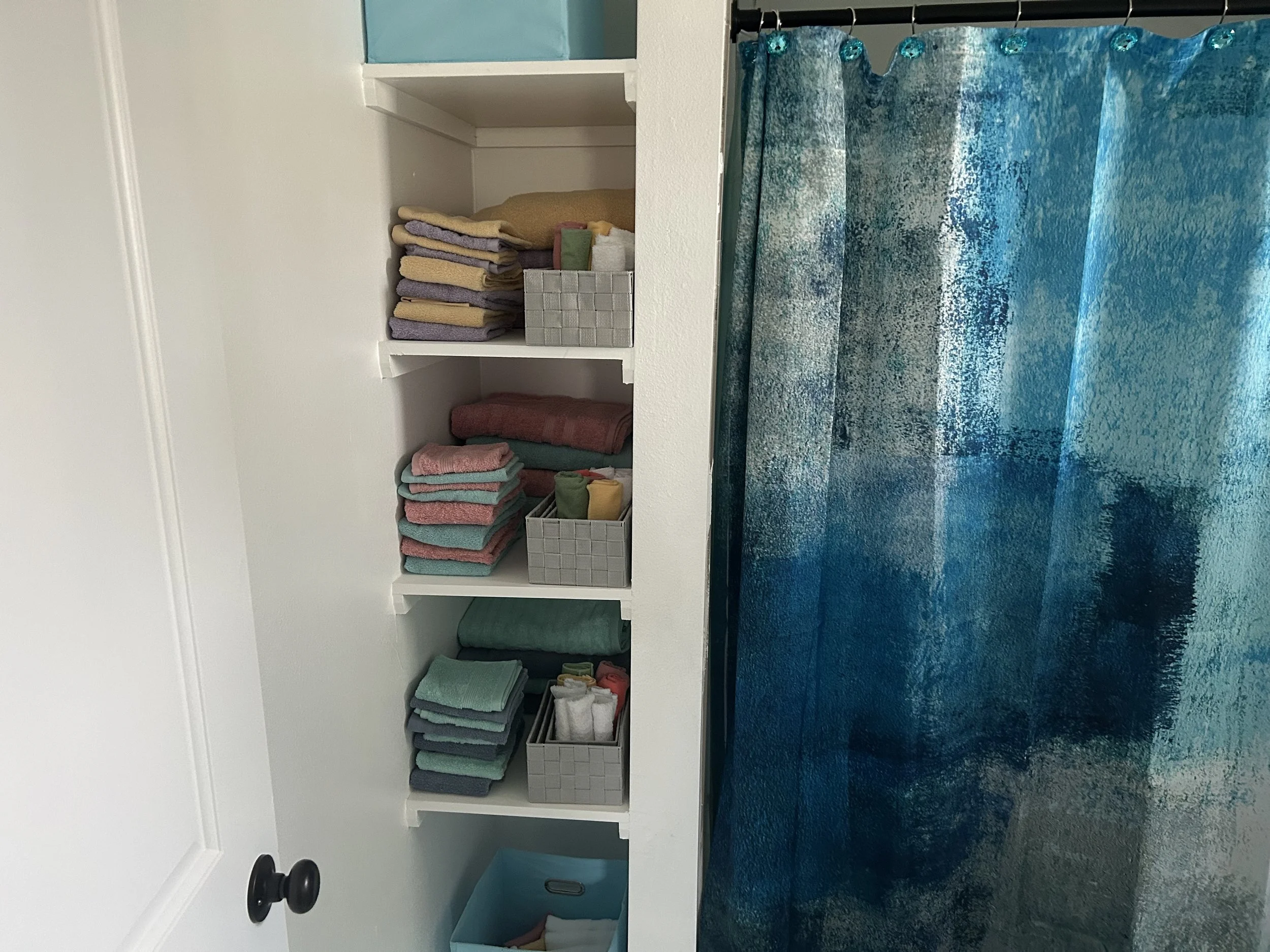 A white shelf with neatly folded towels in pastel colors, a woven basket, and a few small items, positioned against a light-colored wall. To the right, a blue and white textured shower curtain hangs, partially covering the shower or bathtub area.
