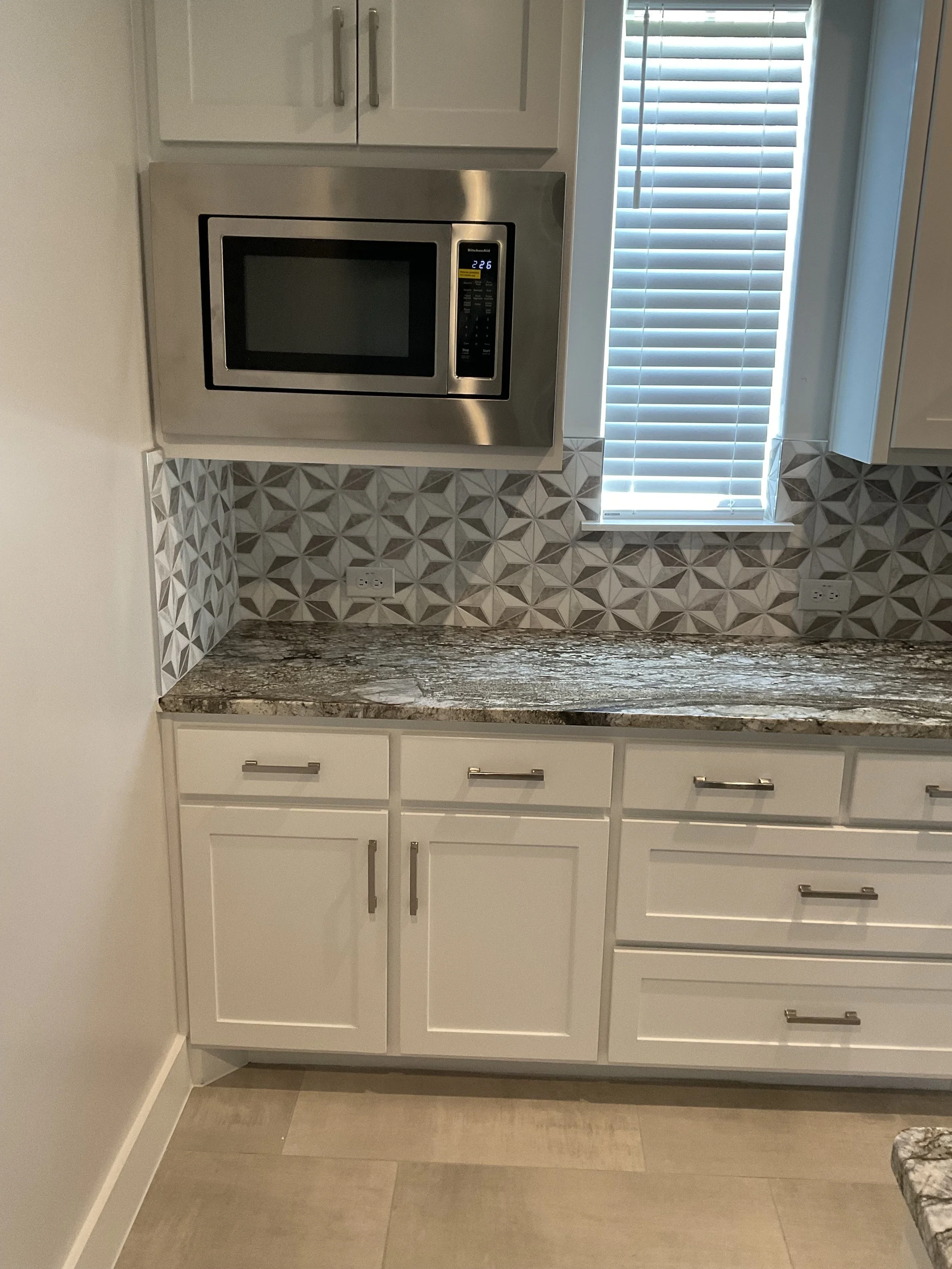 A modern kitchen with white cabinets, granite countertop, patterned tile backsplash, microwave oven, and window with blinds.