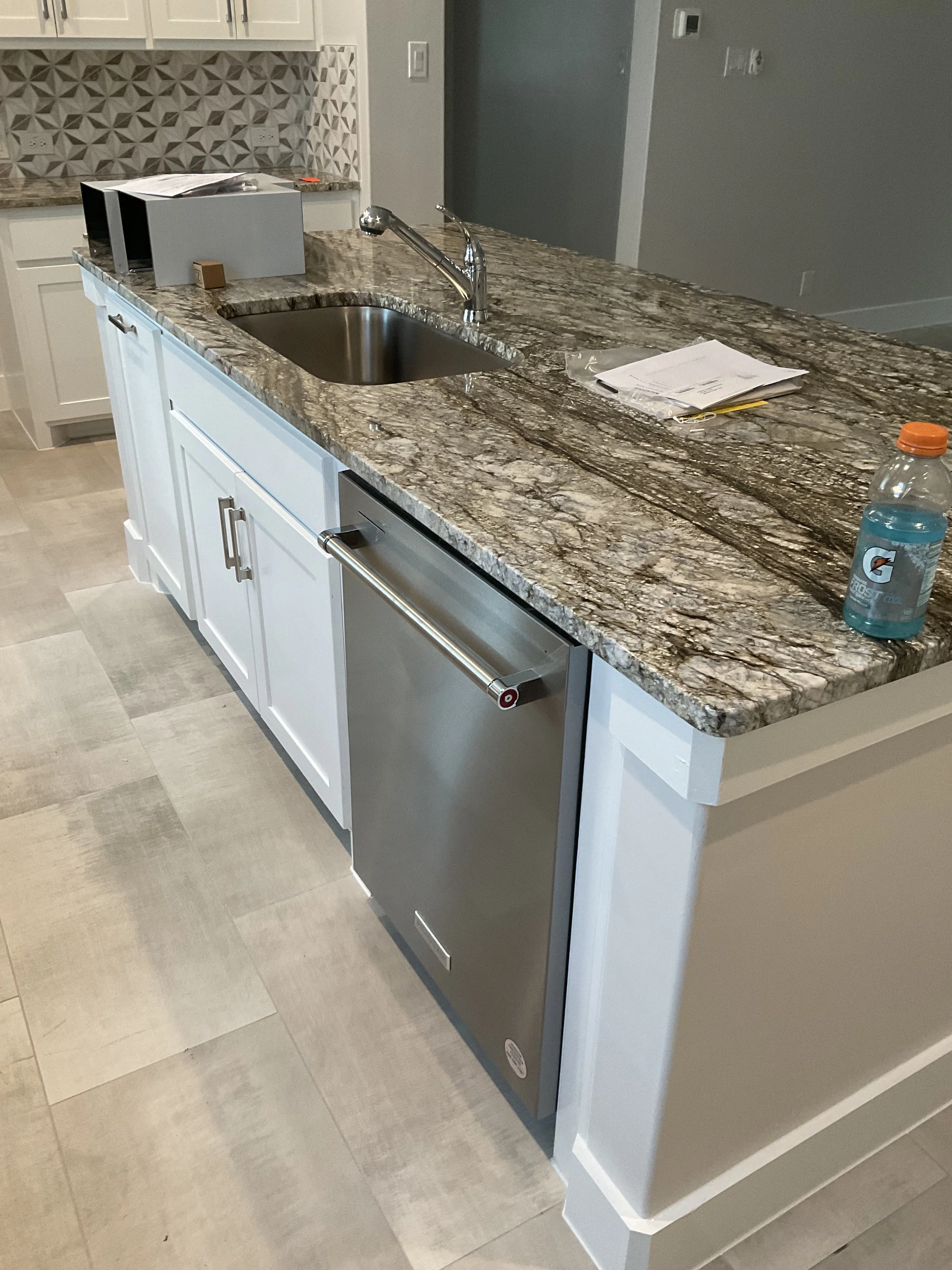 The image shows a kitchen island with a granite countertop, a sink, a faucet, some papers, a bottle of Gatorade, and a few other items. The background includes white cabinets and a decorative backsplash.