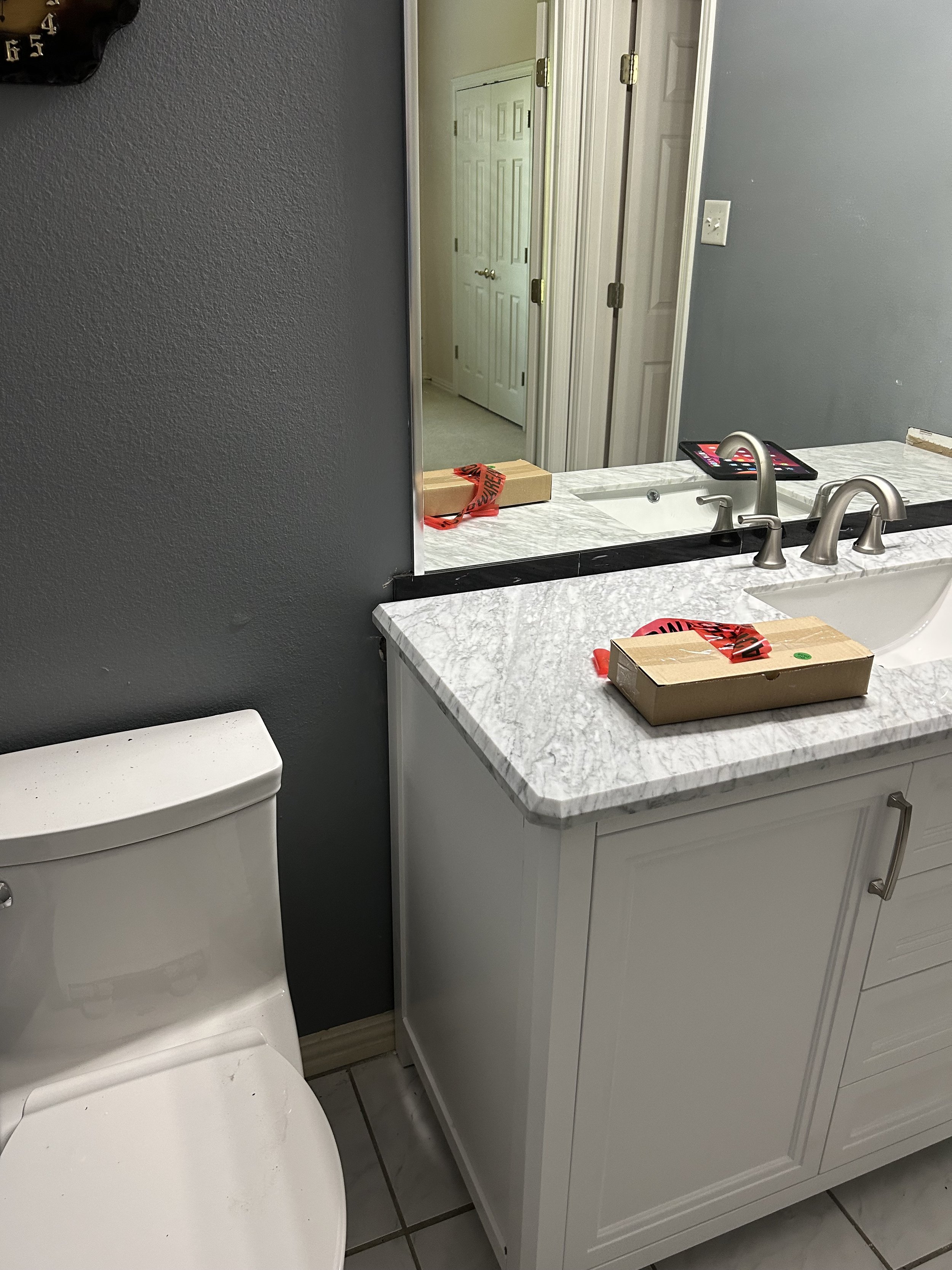 A bathroom with a toilet, a marble countertop with a sink, and a setting to the right for a mirror. There are paper towels on top of the counter, a cardboard box, and a tablet device. The mirror reflects a hallway with white double doors and an addit
