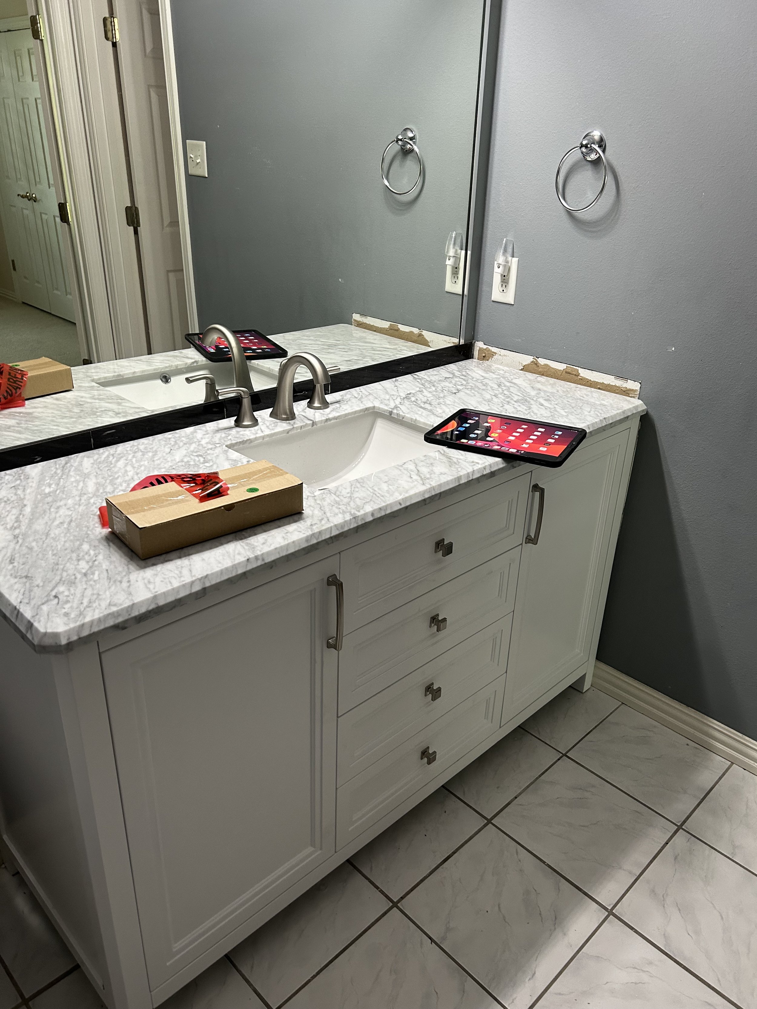 Bathroom vanity with a marble countertop, two sinks, and a large mirror. Two cardboard boxes with red tape are on the counter. A tablet lies on the counter. The wall has a towel ring and electrical outlets. The floor has large, white marble tiles wit