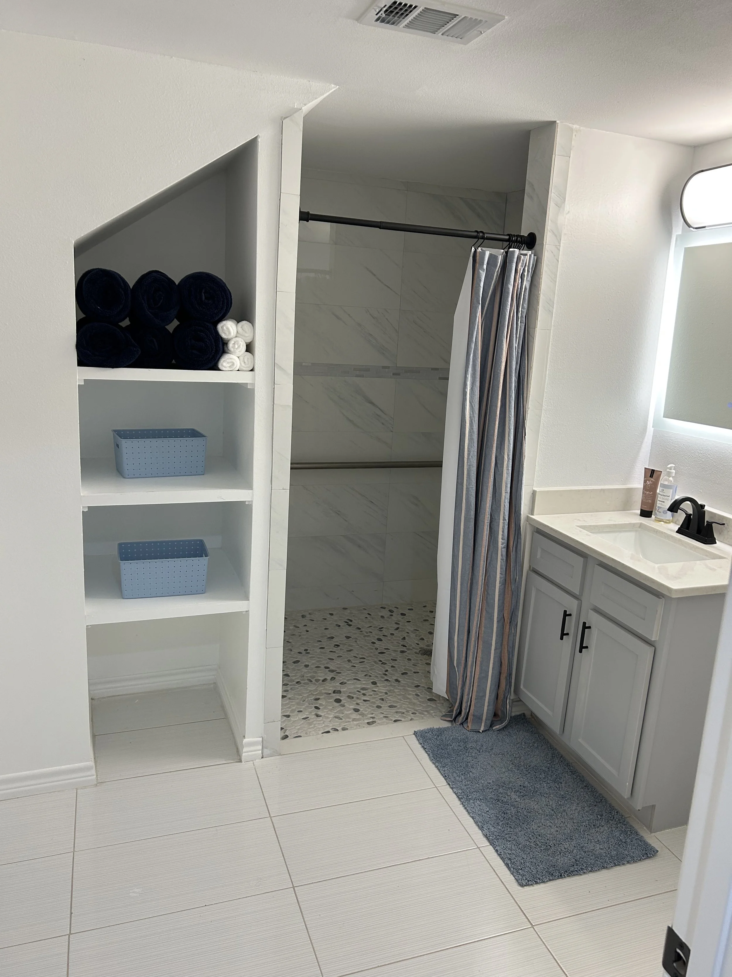 View of a bathroom with a shower area, partially covered by a striped shower curtain. To the right, there is a white vanity with a sink, faucet, and toiletries. To the left, built-in shelves hold rolled towels and blue storage baskets. The flooring c