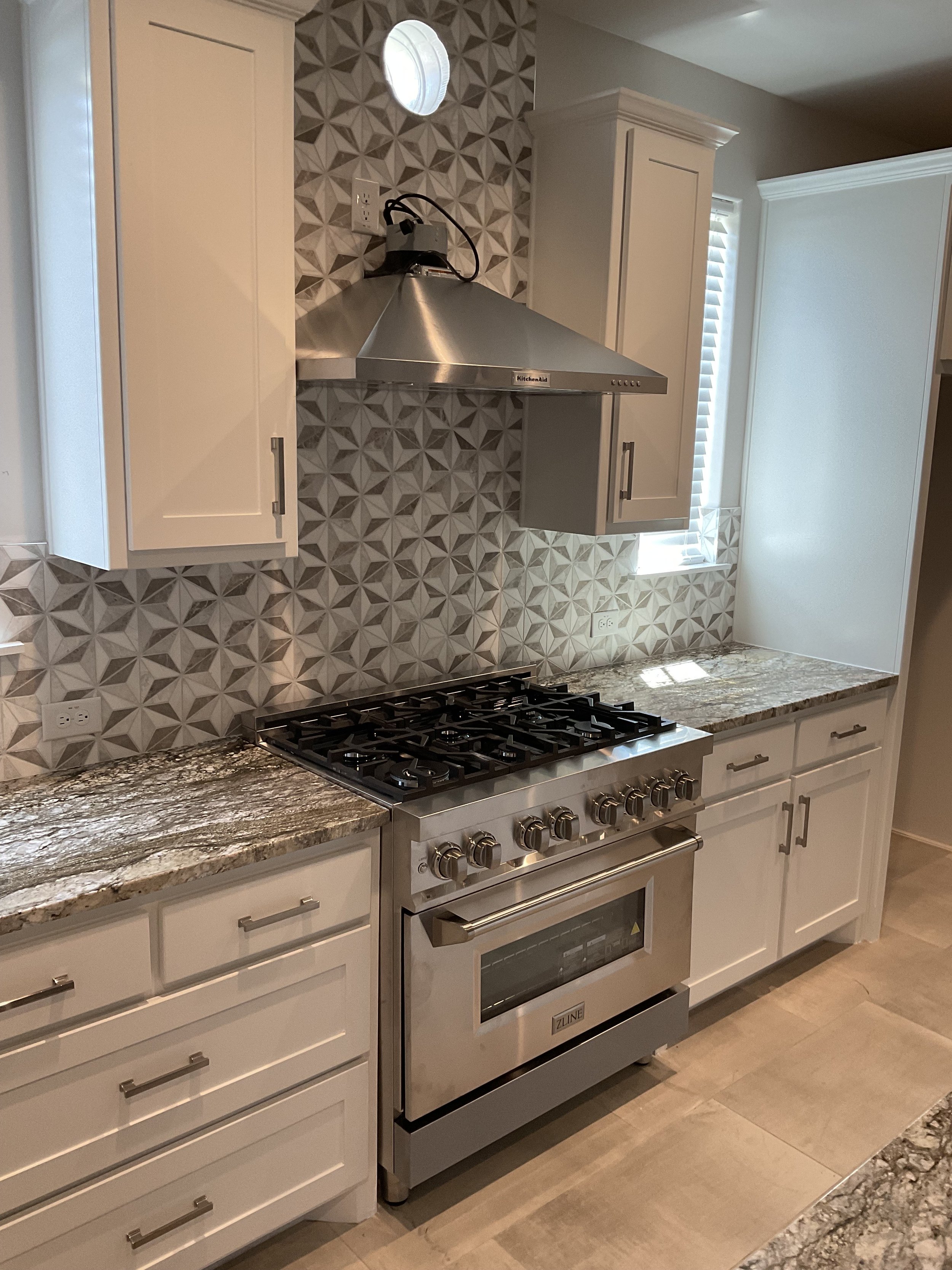 Modern kitchen with granite countertops, white cabinets, and a stainless steel stove and range hood. Patterned backsplash above the stove, small window with blinds to the right.