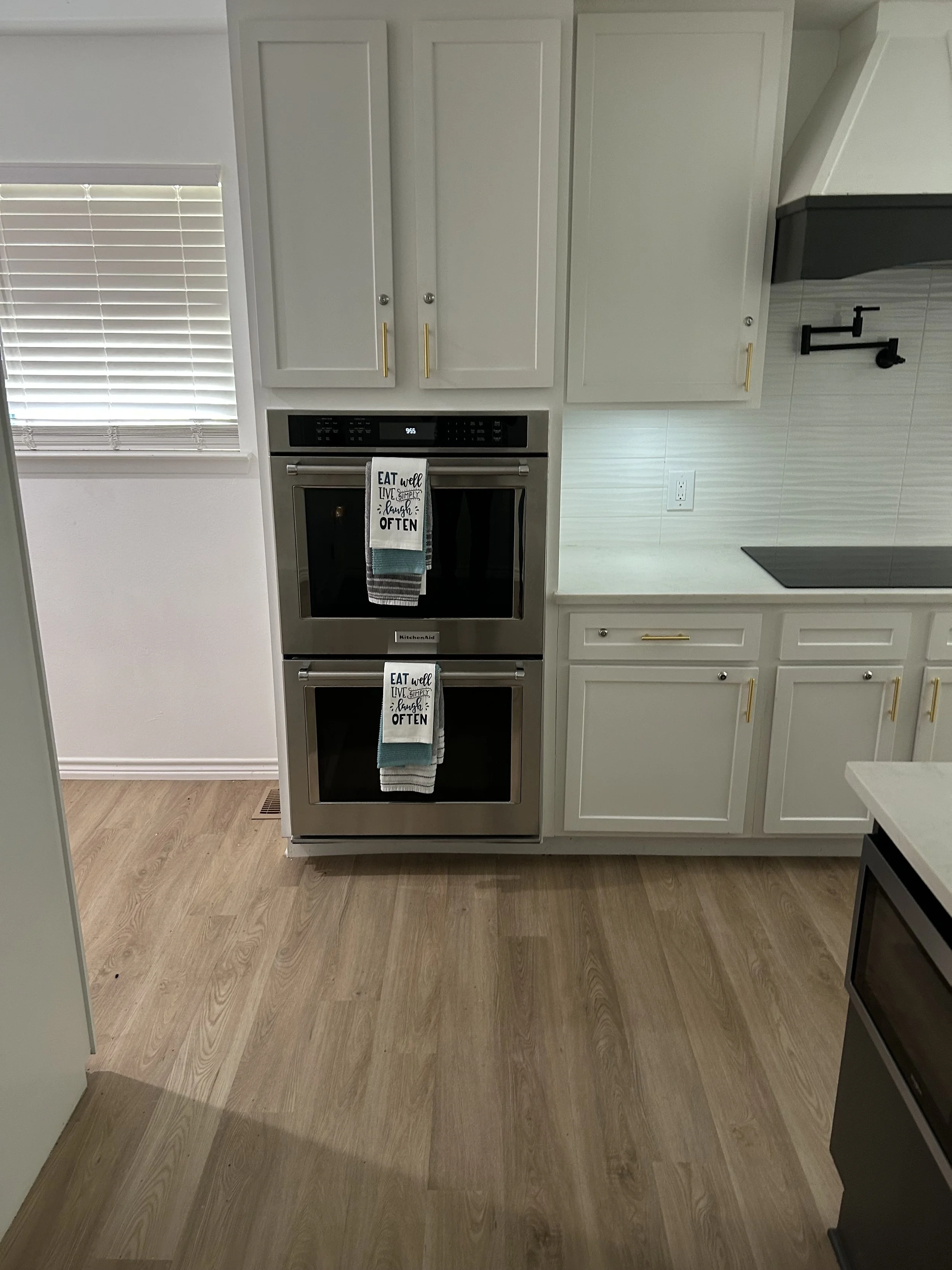 A kitchen with white cabinets, a stainless steel double oven, a black faucet, and light wood flooring.