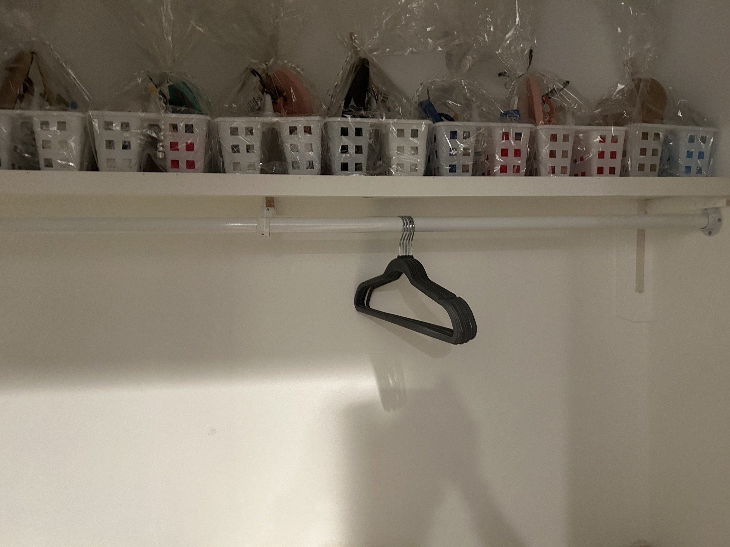 A closet shelf with white plastic bins containing small items wrapped in plastic, with a black clothing hanger hanging from a rail below the shelf.