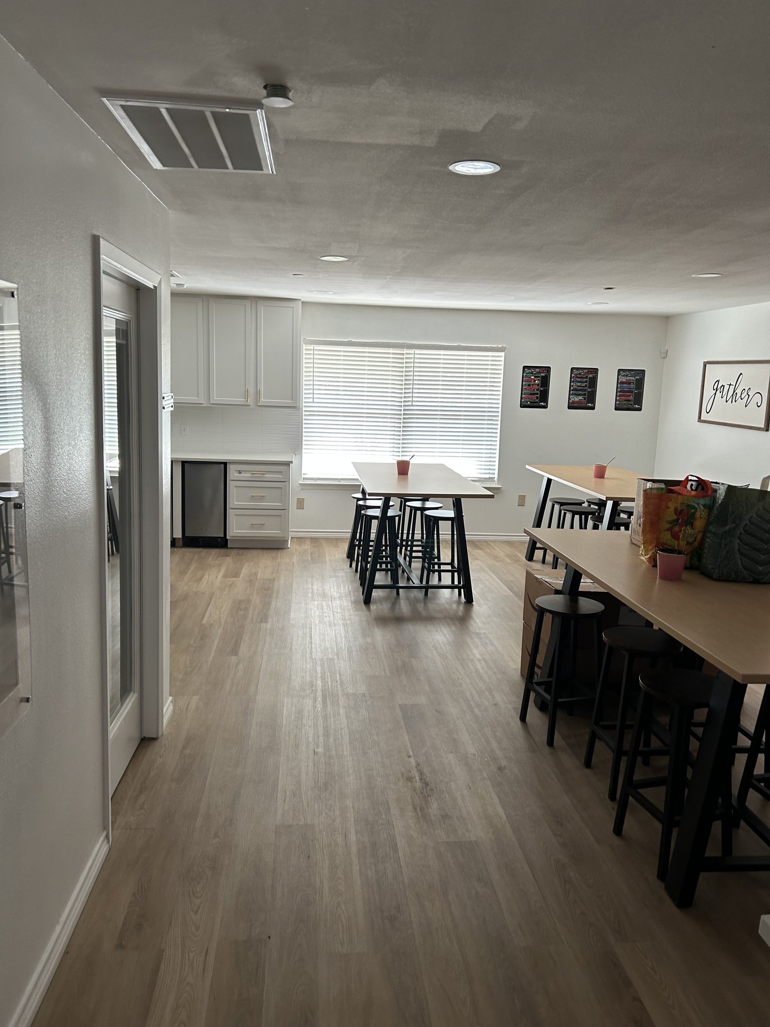 A bright, modern kitchen and dining area with light wood flooring, white cabinets, a large window with blinds, and black stools around high tables with pink cups on top.