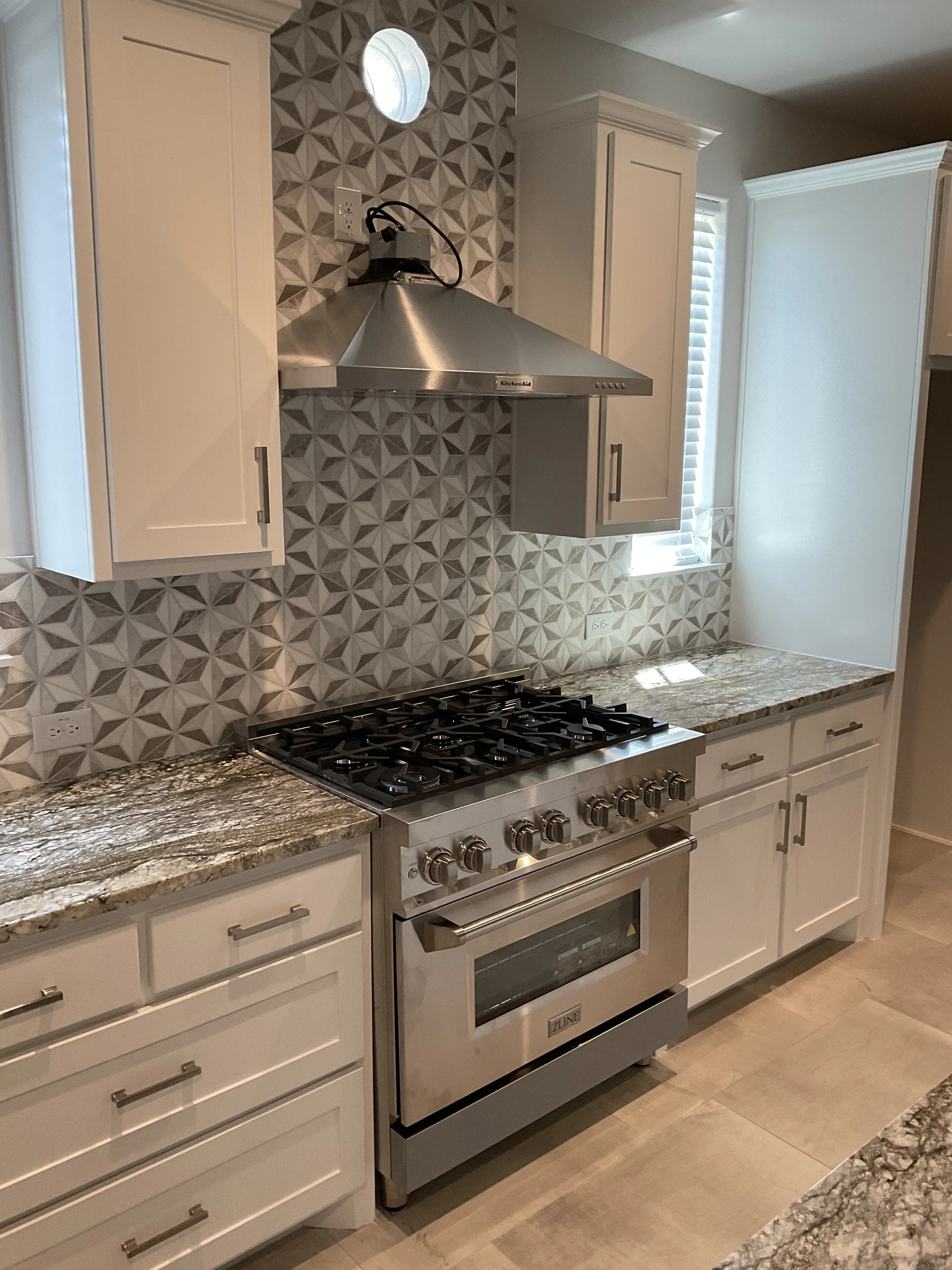 Kitchen with white cabinets, granite countertop, stainless steel stove and oven, stainless steel range hood, geometric patterned backsplash, and a small window with blinds.