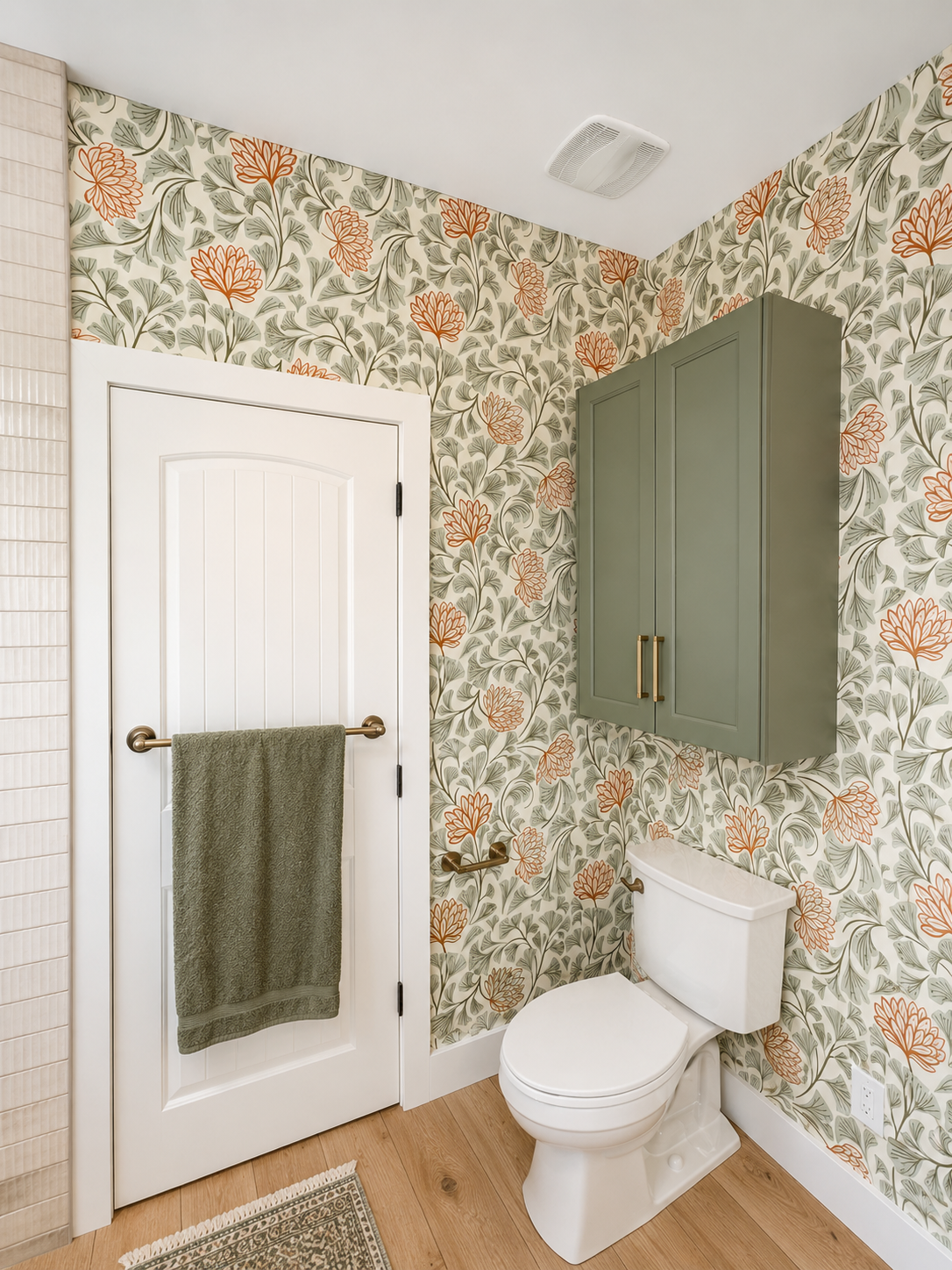 A bathroom with floral wallpaper, a white toilet, a green wall cabinet, a white door with a towel bar, and wood flooring.