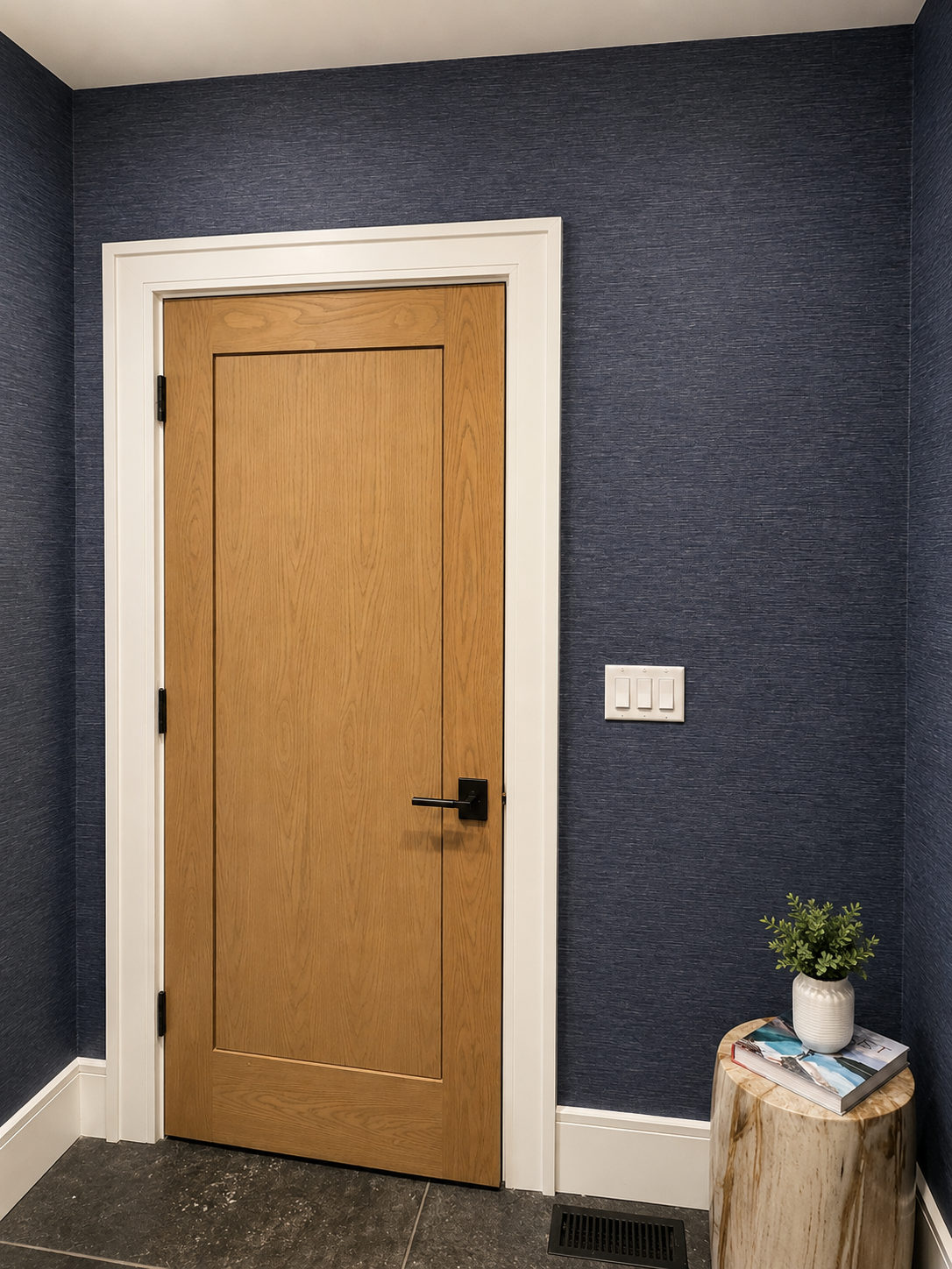 Interior wall with blue textured wallpaper, a closed wooden door with black handle, three light switches on the wall, a small wooden side table with a potted plant and magazines, and a floor vent.