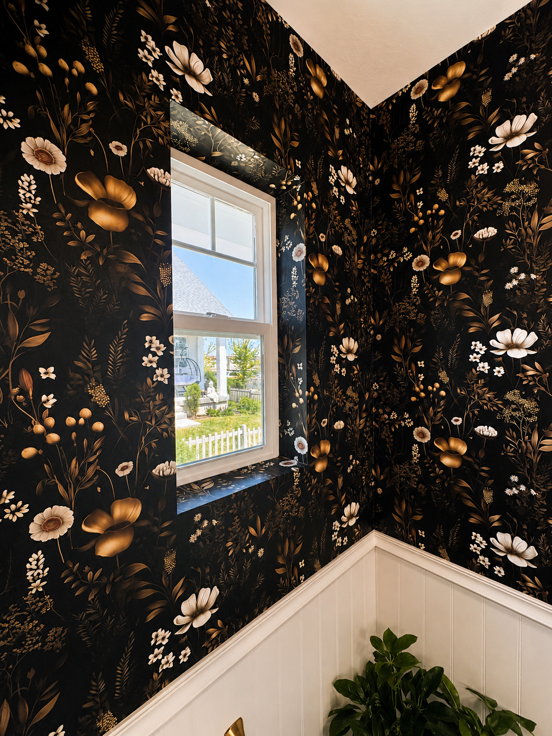 A corner of a room with black floral wallpaper featuring gold and white flowers, a window showing a yard with a white picket fence, greenery, and a house outside, and a green indoor plant in the corner.