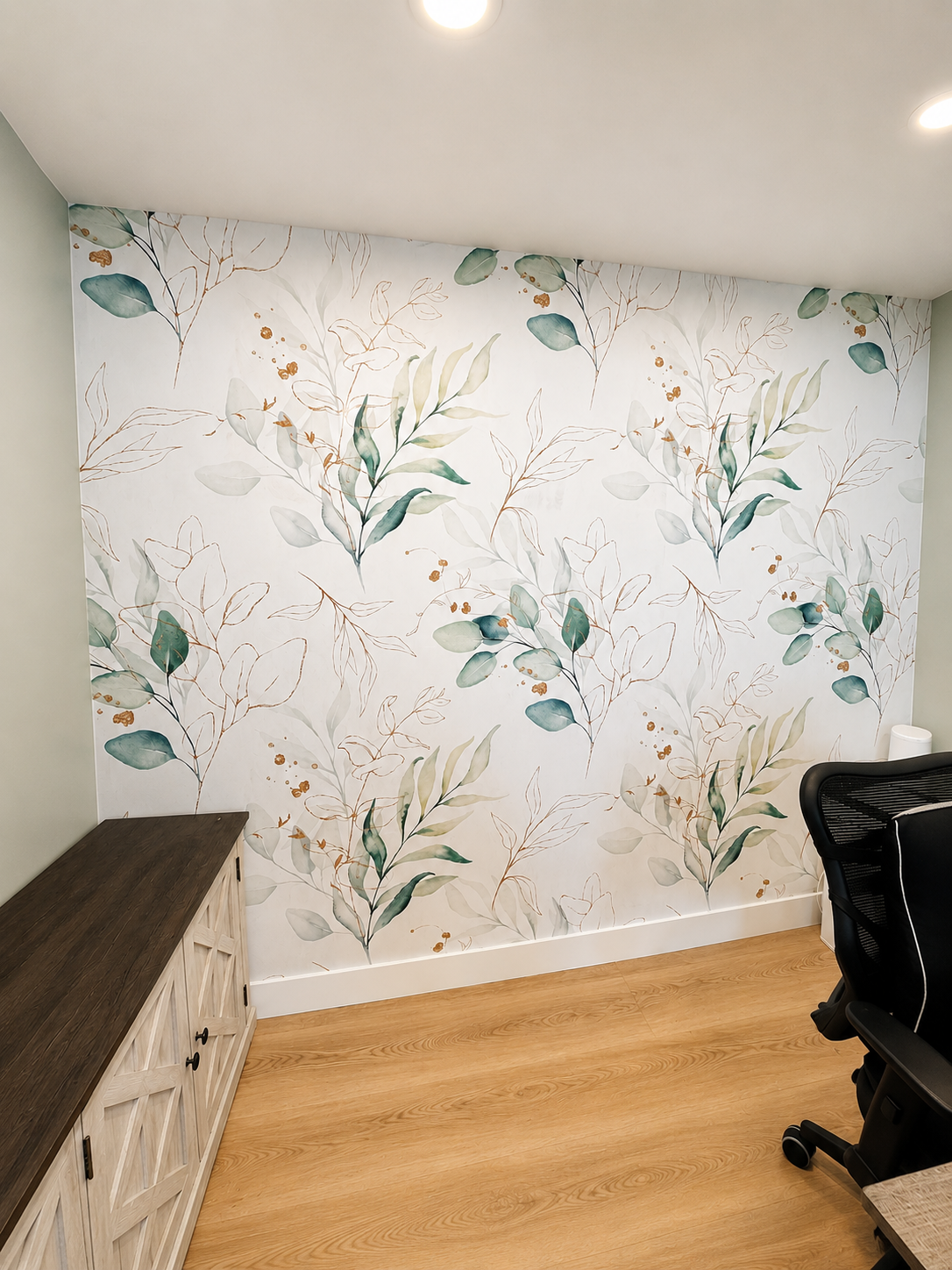 A wall with light-colored floral wallpaper featuring green and beige leaves and small orange accents, adjacent to a wooden cabinet and a black office chair with a wooden base on a light wood floor.