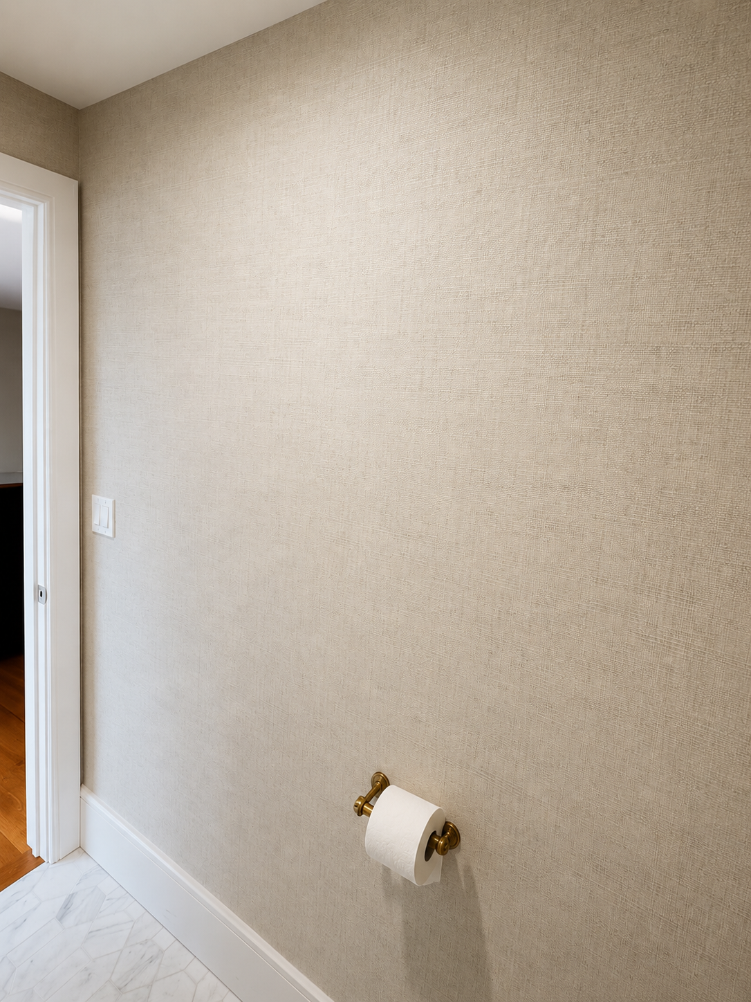 Interior beige textured wallpaper with a white baseboard, a white door frame, and a toilet paper roll mounted on a brass holder.