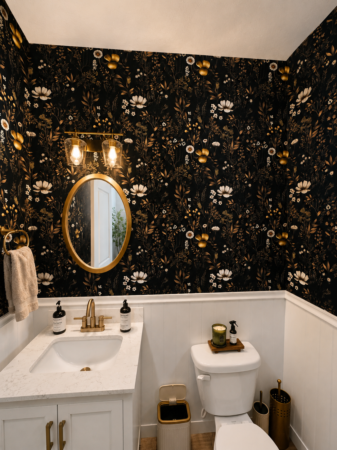 Bathroom with black floral wallpaper, oval mirror with gold frame, white vanity with gold fixtures, white toilet, gold and black trash bin, and gold accents on accessories.