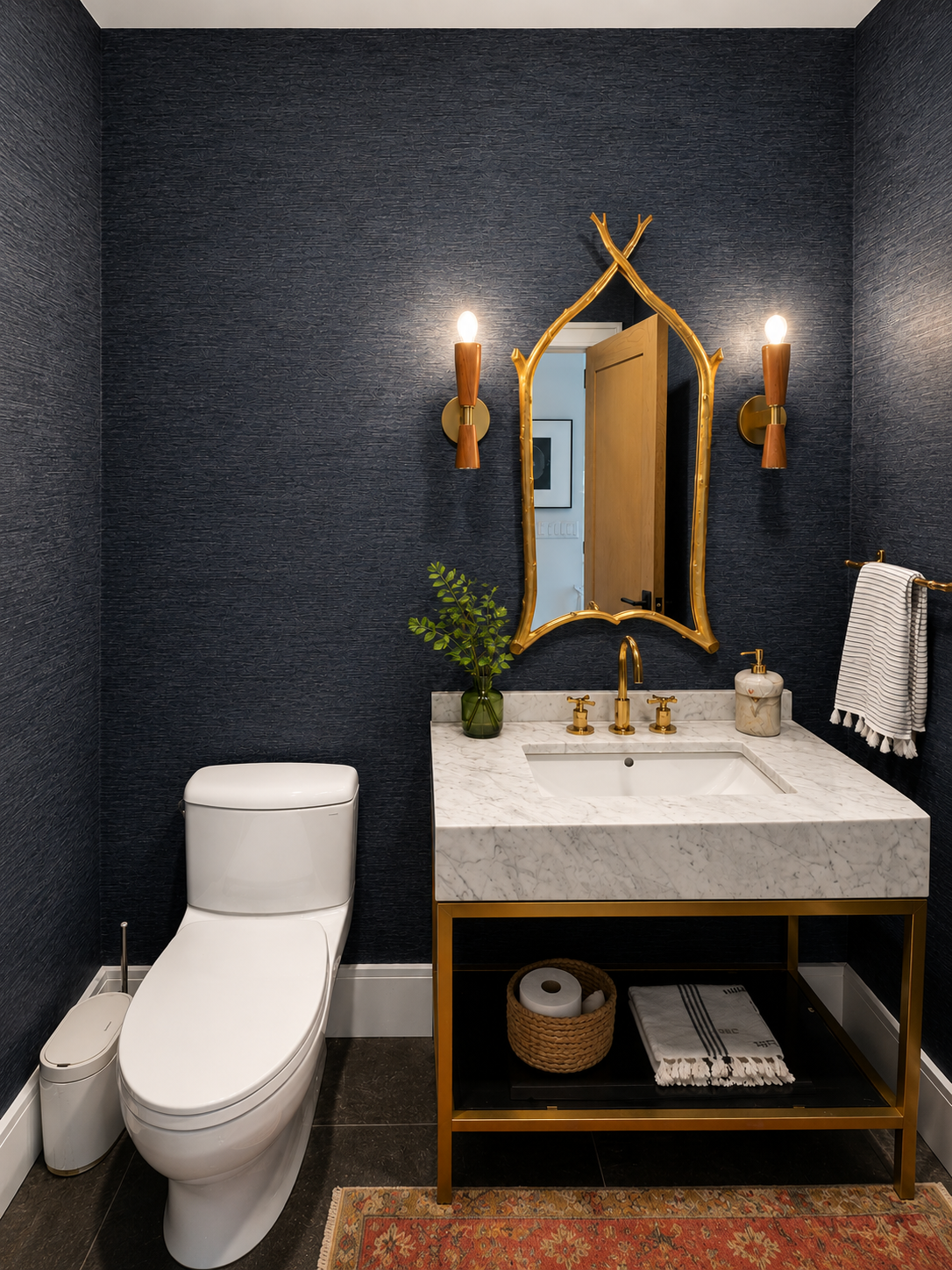 A  with dark textured, gold accents, and a marble sink vanity. It features a decorative mirror, two wall sconces, a small green plant, and a towel hanging on a gold rod. There's a white toilet, a matching waste bin, and a woven basket with toilet pap