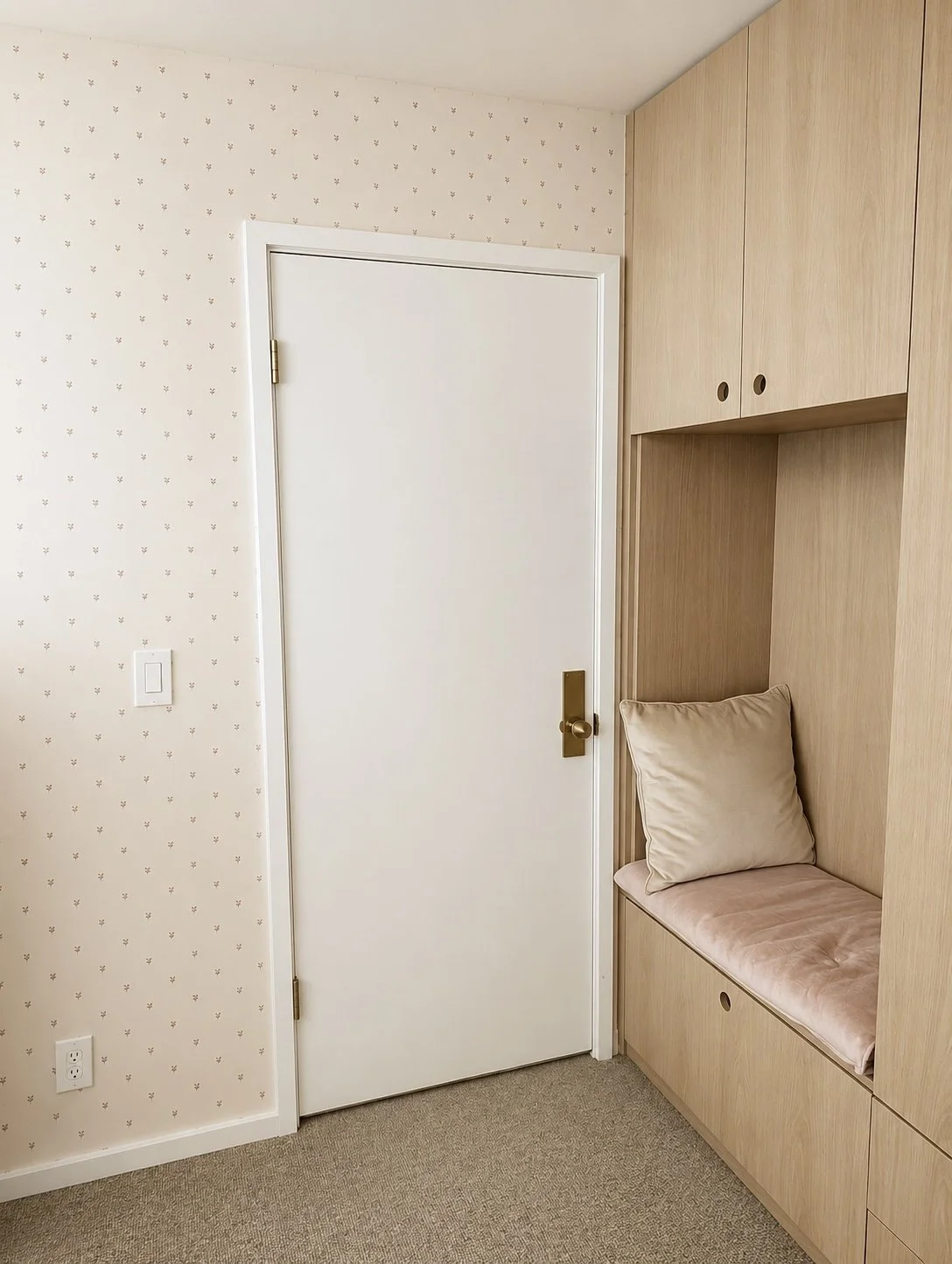 A cozy corner nook with blush wallpaper with a white door, wooden storage cabinets, and a built-in bench with pink cushioning and a beige pillow.