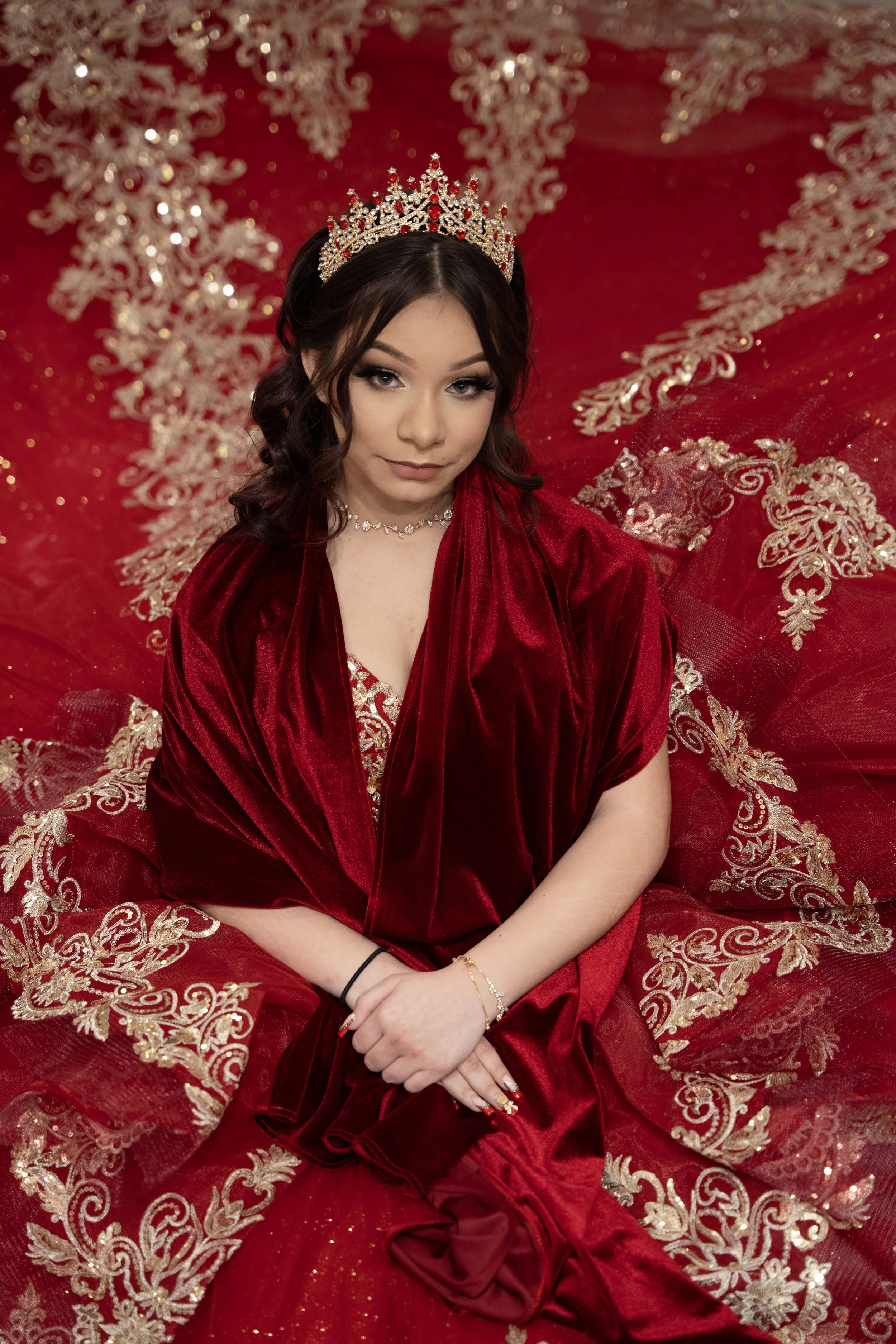 A young woman in a red velvet gown with gold embroidery, wearing a rhinestone tiara and jewelry, sitting on a red and gold embroidered fabric background.
