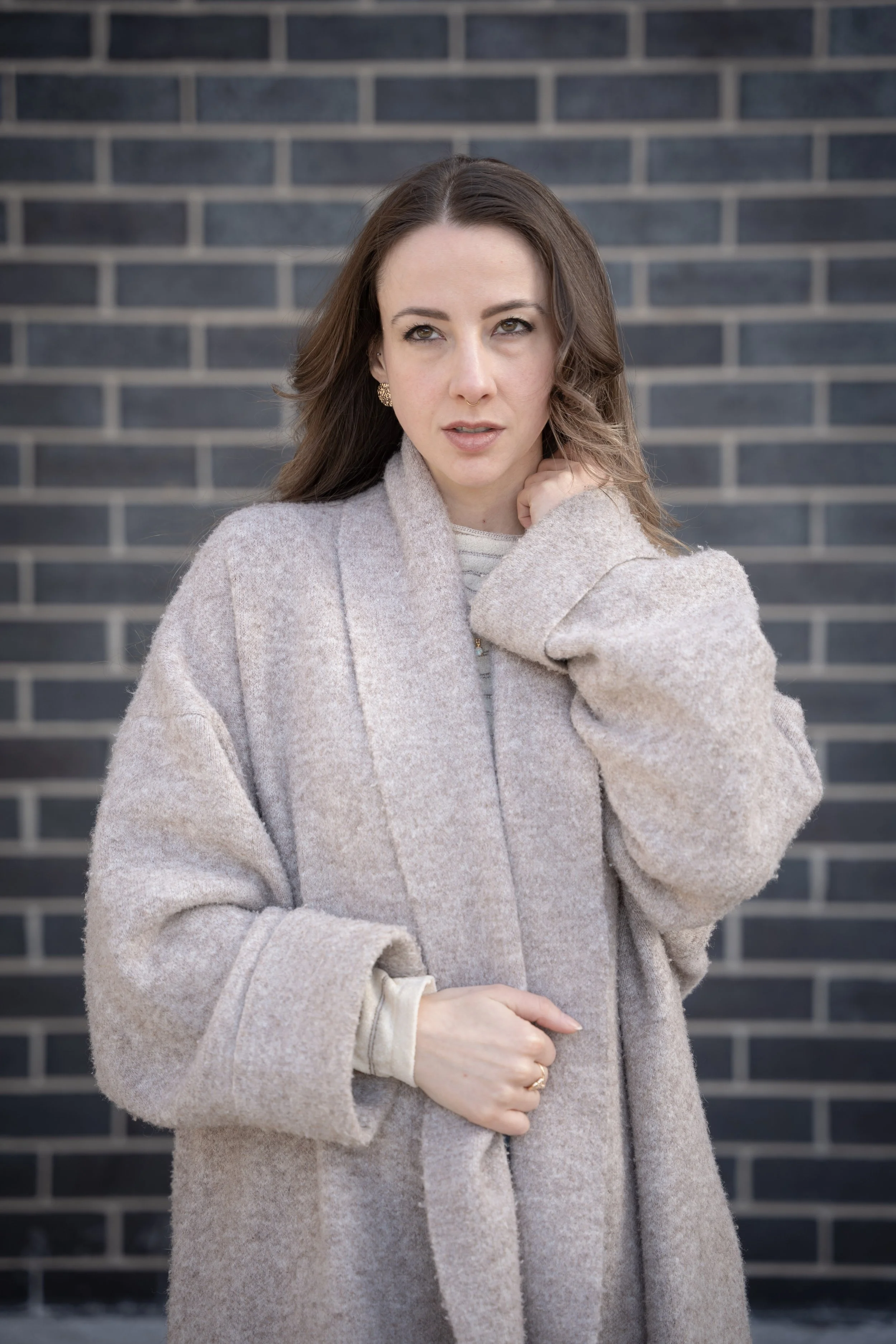 A woman with long brown hair standing outdoors in front of a dark brick wall, wearing a beige wool coat and a striped shirt underneath.