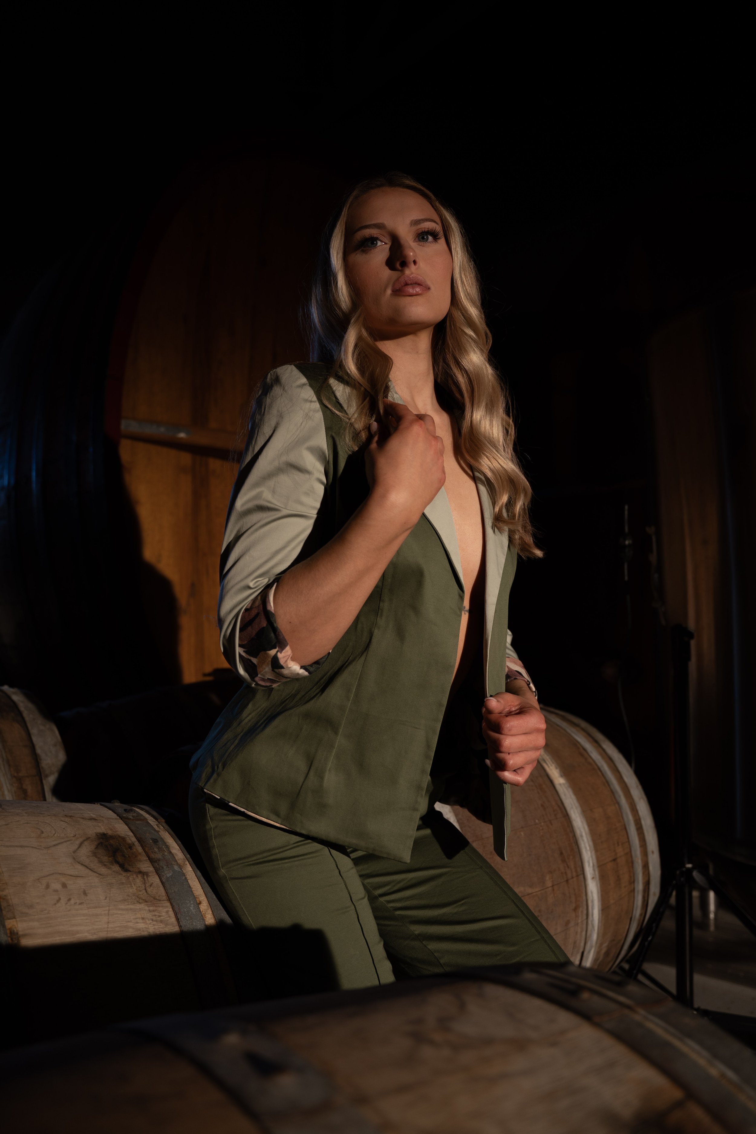 A woman with long blonde hair wearing a partially unzipped green jacket and matching pants, sitting on a wooden wine barrel in a dimly lit room with wooden walls and barrels in the background.