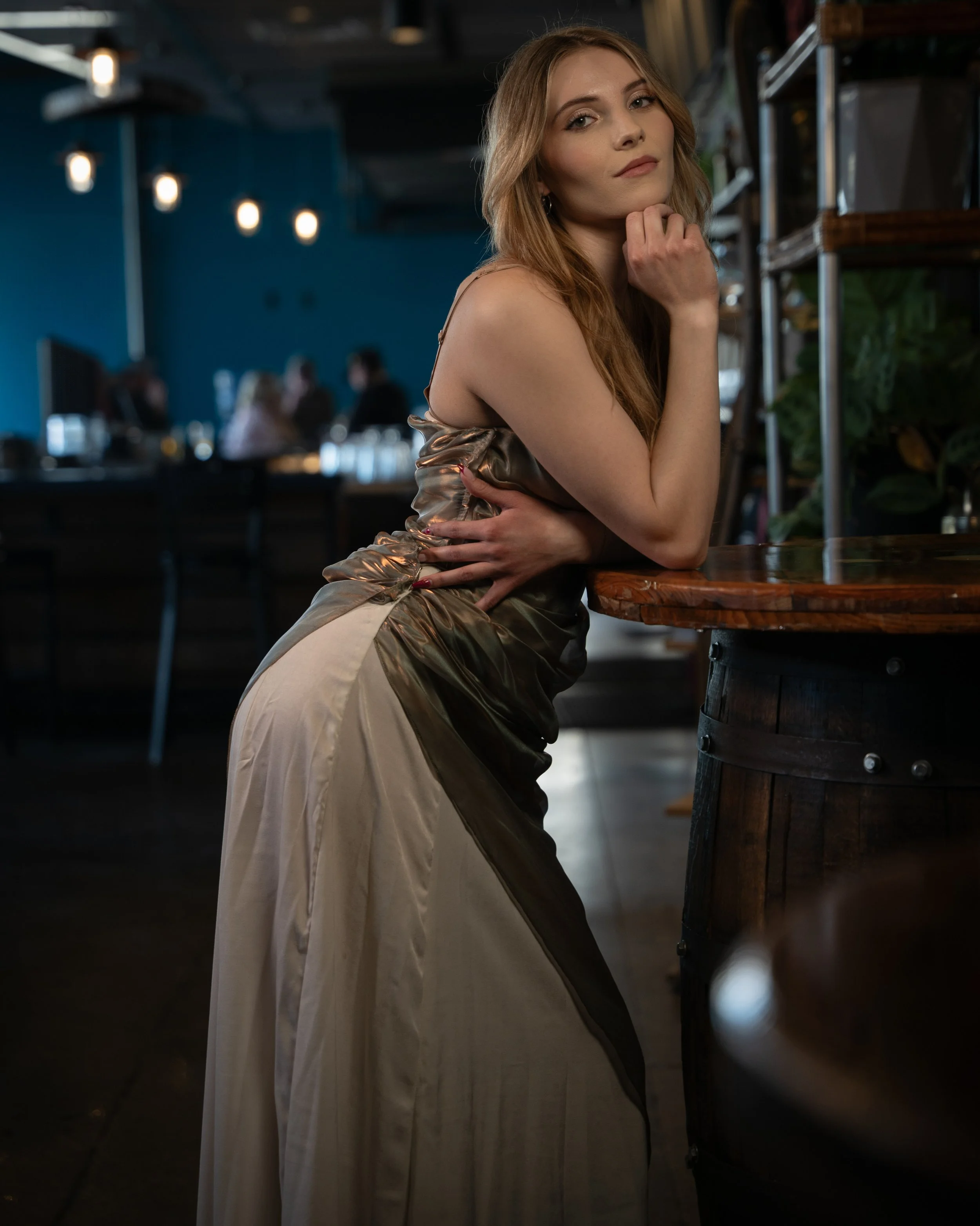 A woman with long, light brown hair posing in a restaurant or bar, leaning on a round wooden table, wearing a satin dress with asymmetrical design, looking at the camera with a confident expression.