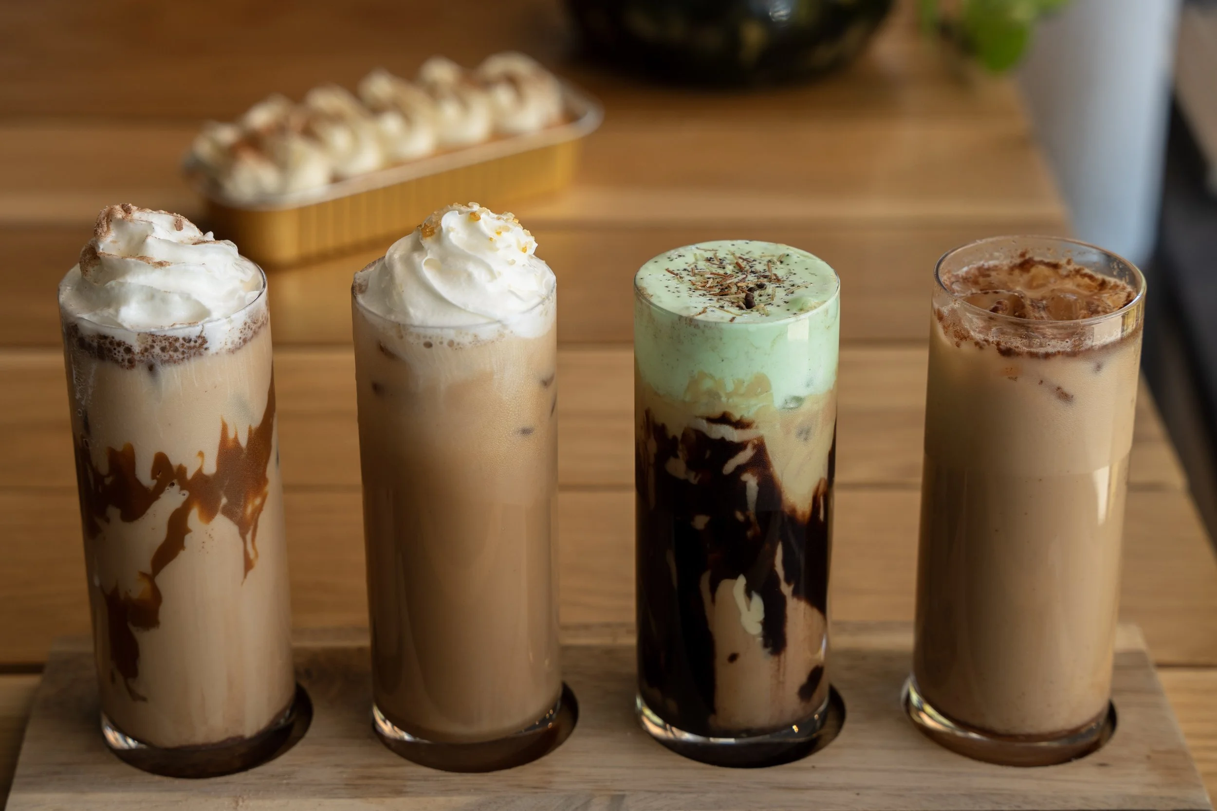 Four tall glasses of iced coffee drinks with whipped cream and various toppings on a wooden tray, with a tray of small baked goods in the background.