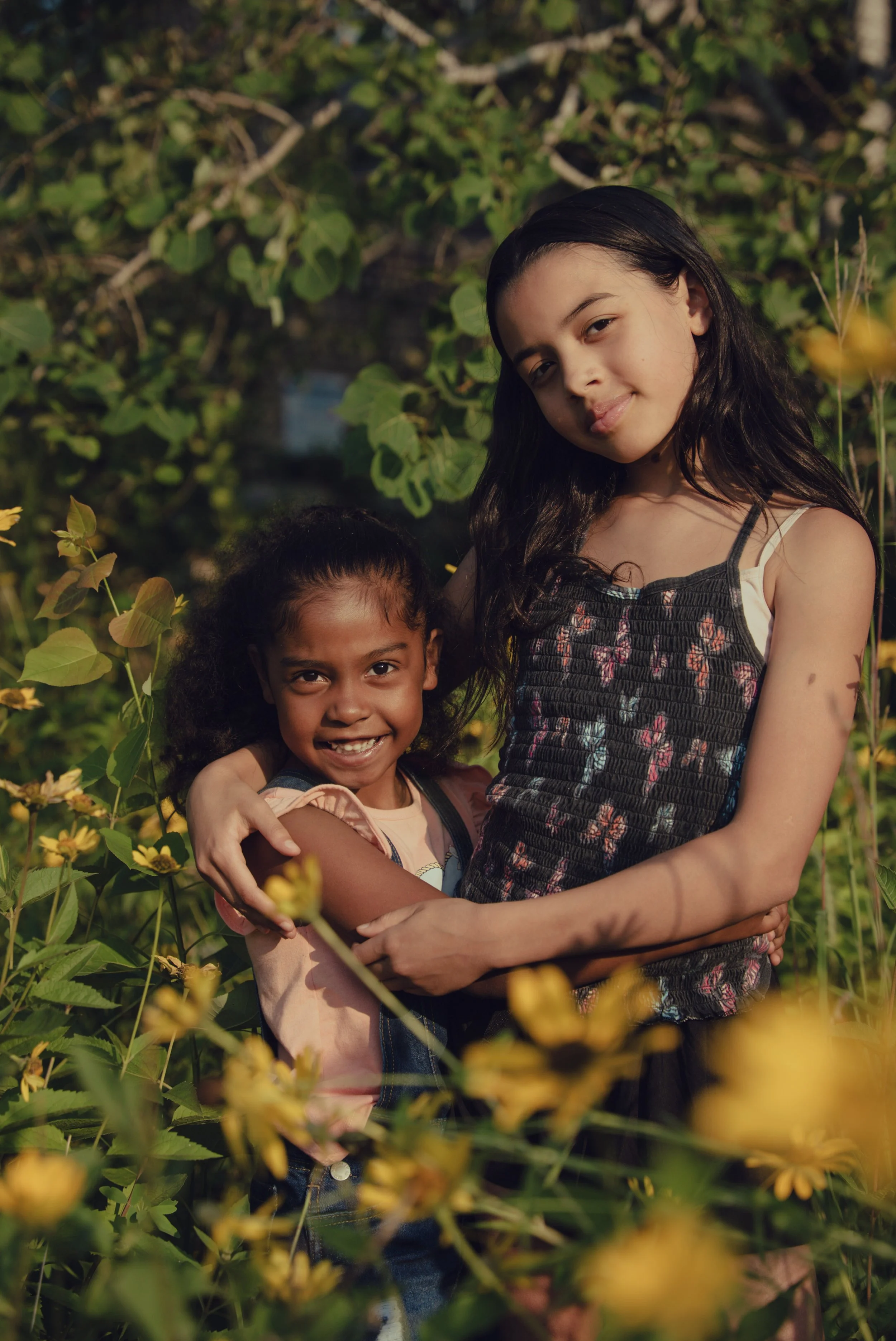 Two young girls, one with dark curly hair and the other with long dark straight hair, smiling and hugging each other among yellow flowers and green foliage during sunlight.