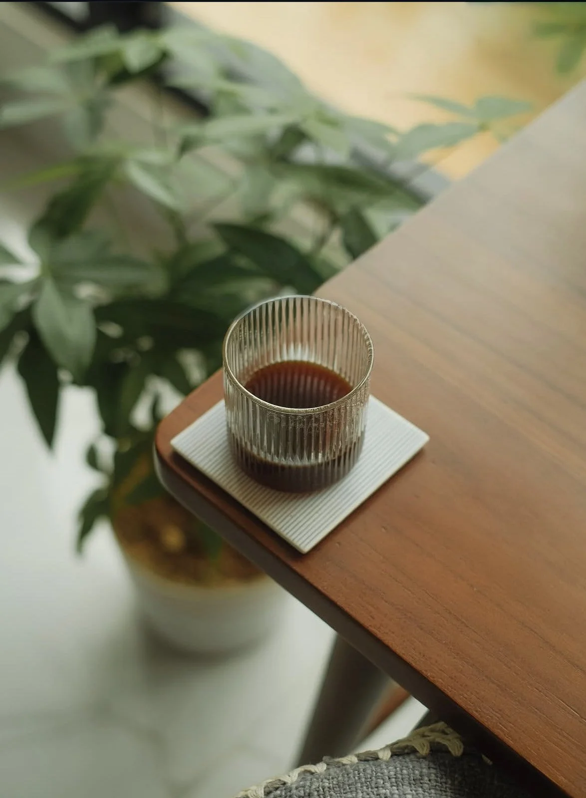 A glass of dark liquid, possibly coffee, on a textured square coaster on a wooden table next to a potted plant with green leaves.