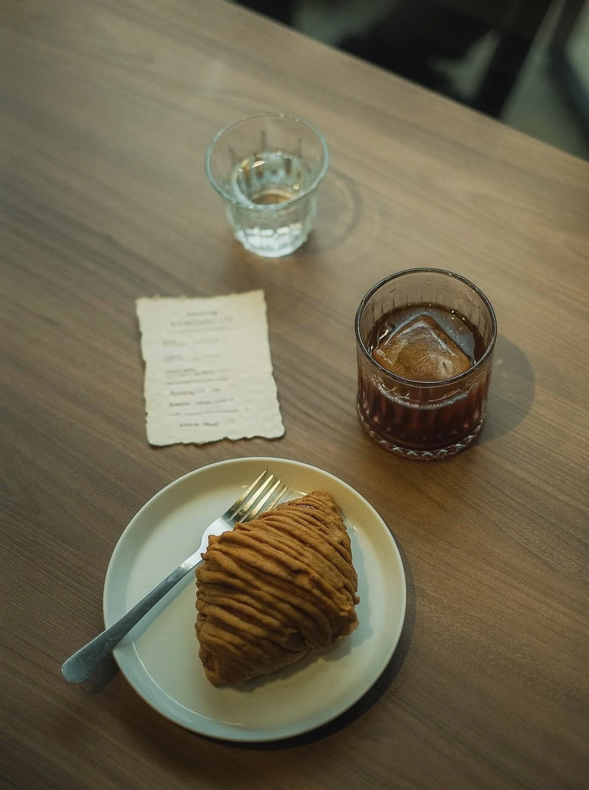 A plate with a golden brown pastry, two glasses with beverages (one with clear liquid and one with dark liquid and ice), a crumpled piece of paper, and a fork on a wooden table.