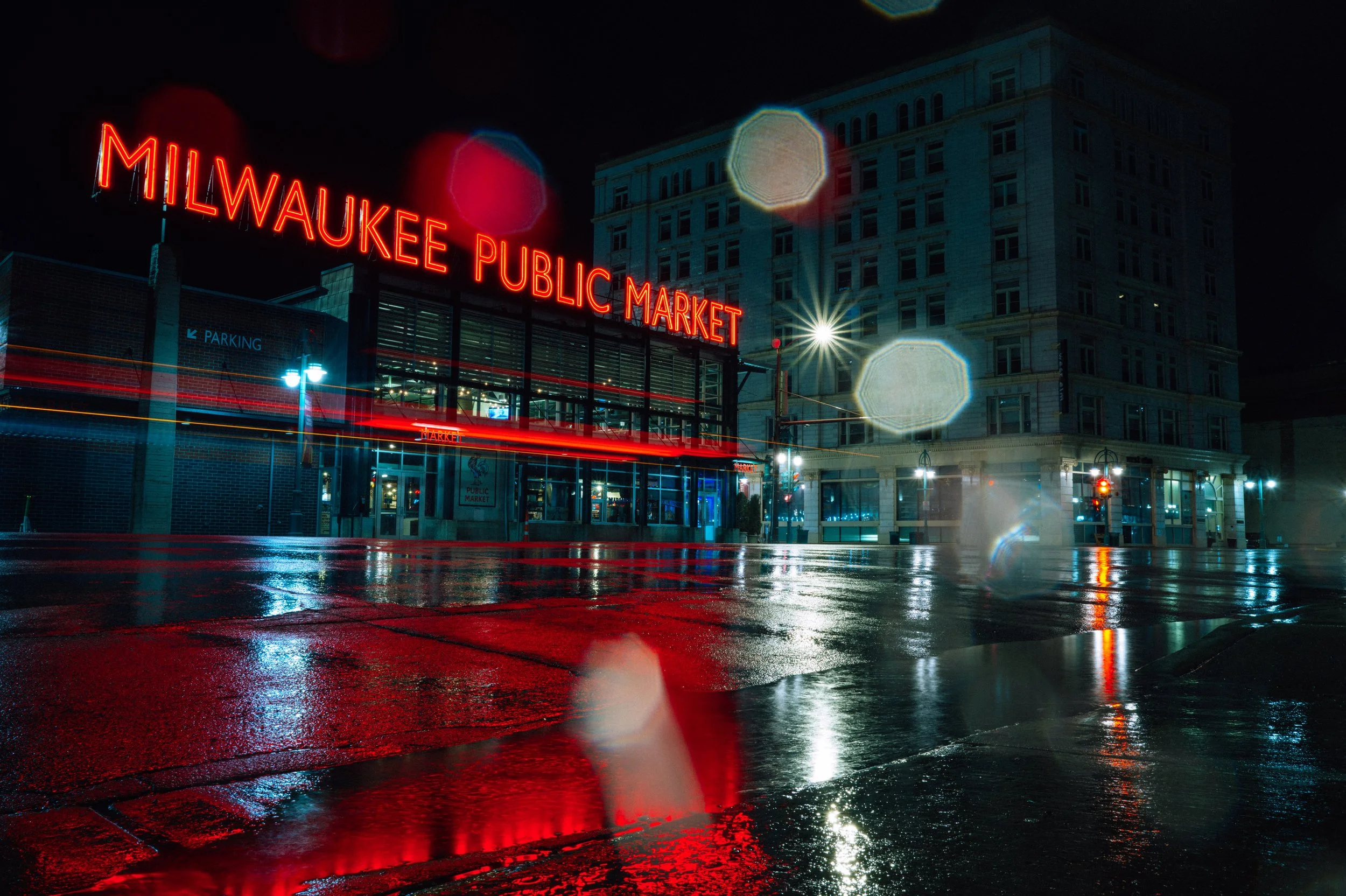 Nighttime view of Milwaukee Public Market with illuminated red neon sign, wet streets reflecting lights, and a multi-story building in the background.