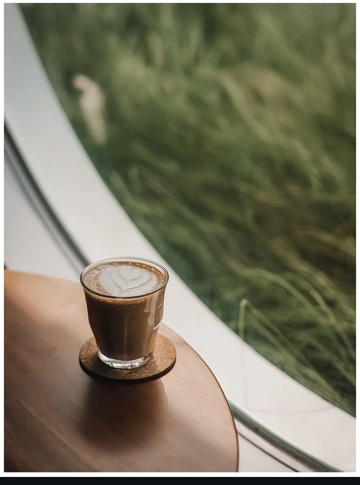 A glass of hot chocolate with foam art in a heart shape on top, placed on a cork coaster on a wooden table near a window with greenery outside.