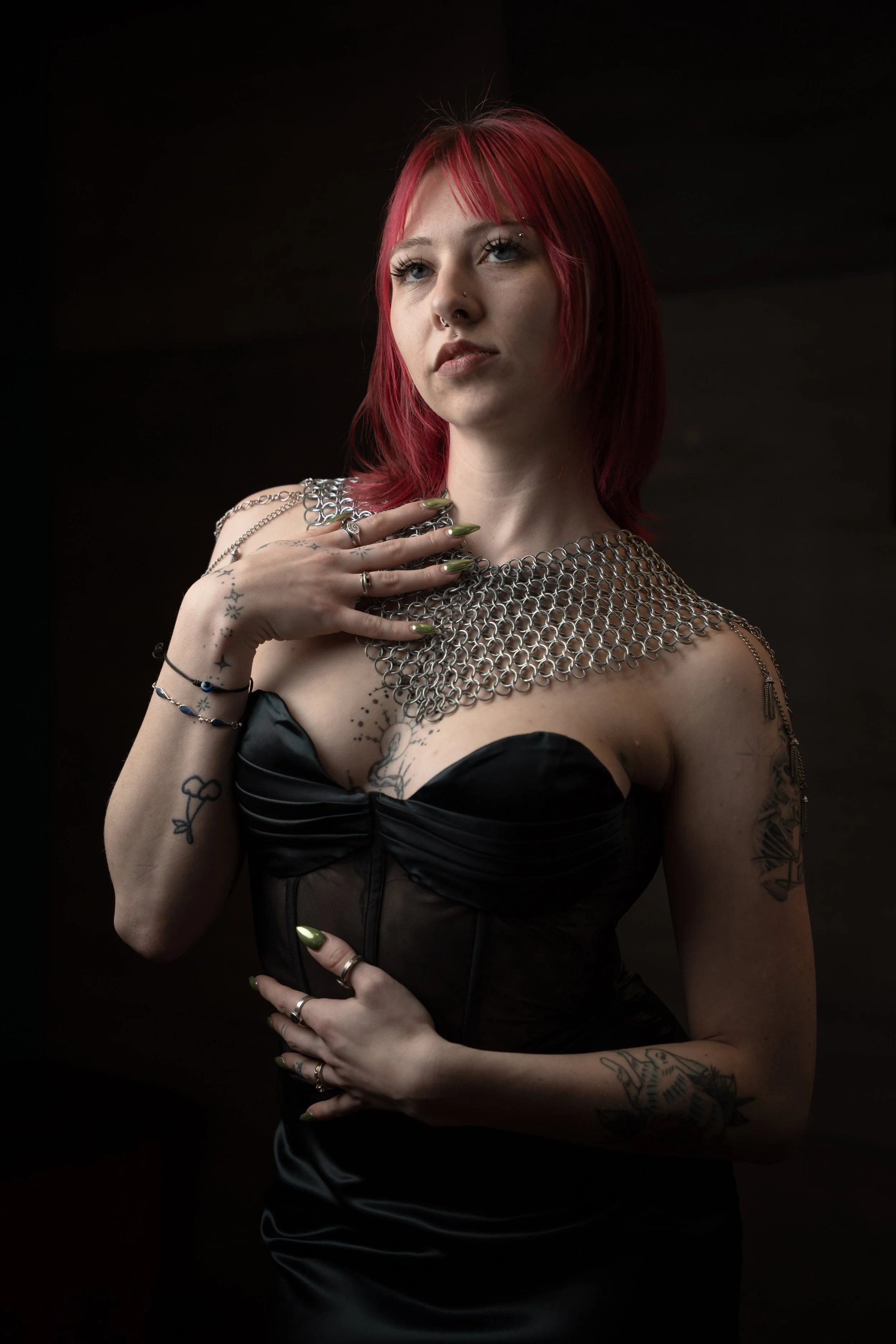 A woman with pink hair and tattoos wearing a black strapless dress and a large chainmail necklace poses against a dark background.