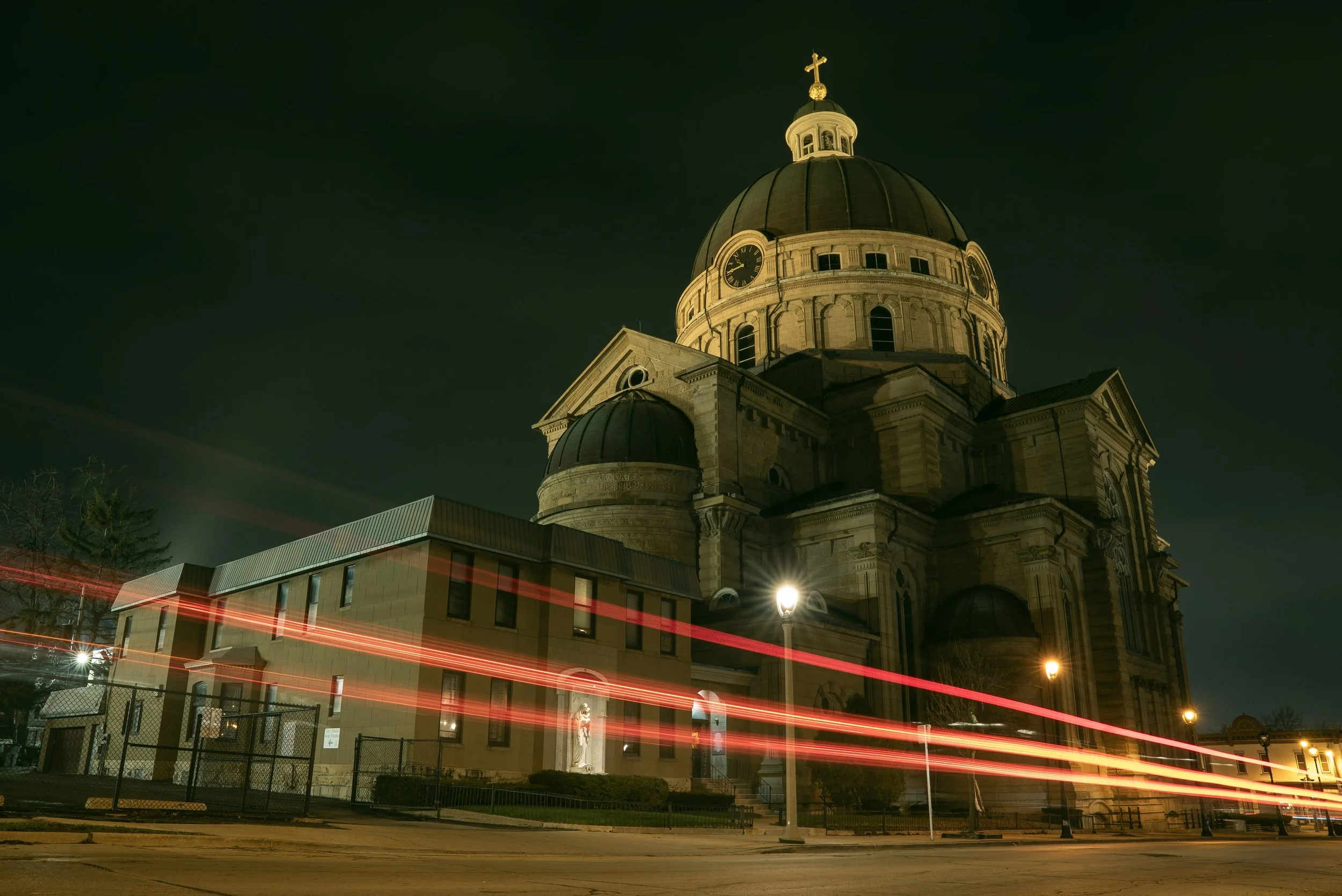 Night view of a large historic church with a prominent dome, clock, and cross on top, illuminated against a dark sky, with light trails from passing cars in the foreground.