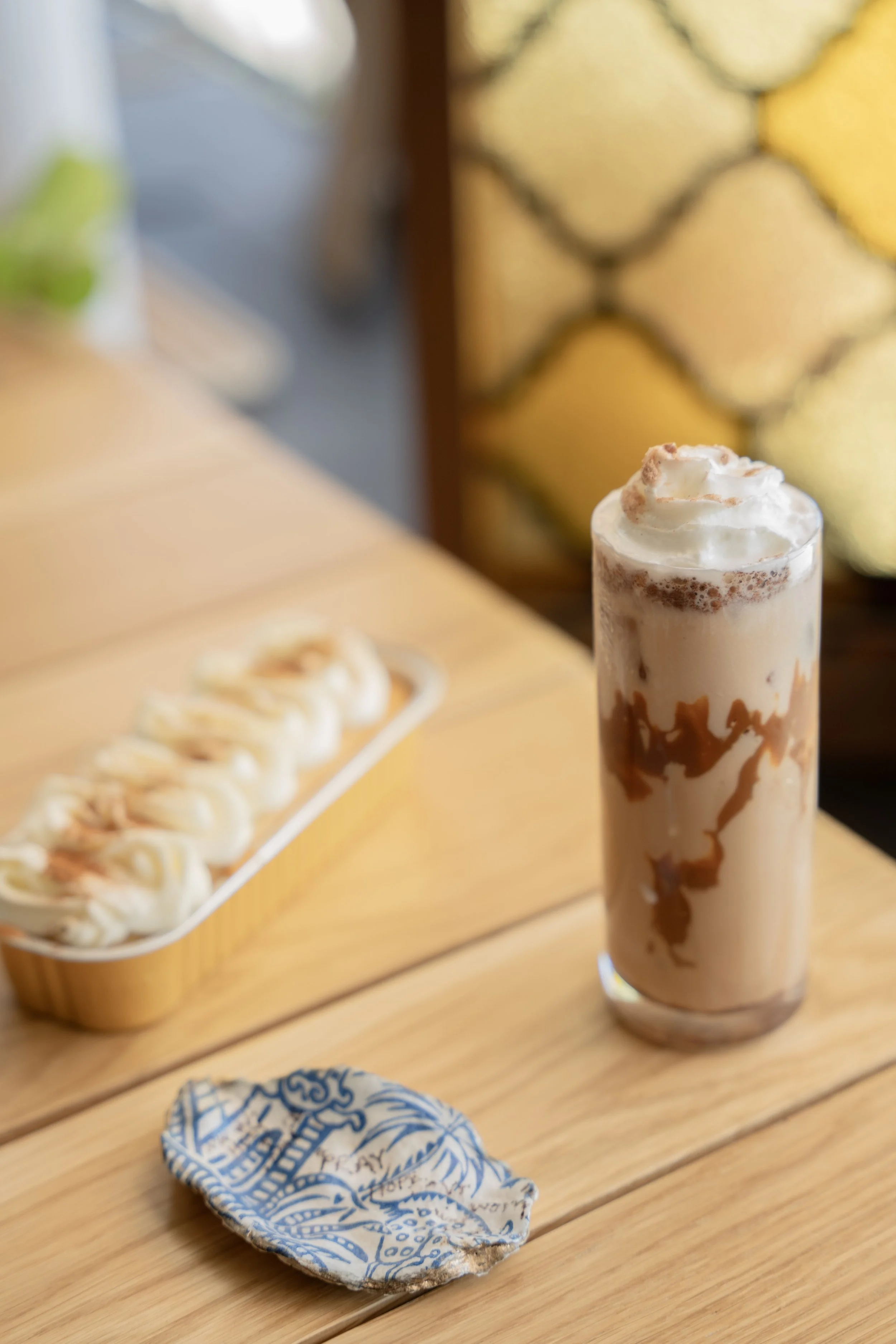 A tall glass of iced coffee topped with whipped cream, with caramel sauce drizzled inside, on a wooden table, alongside a crumpled napkin and a plate of small pastries.