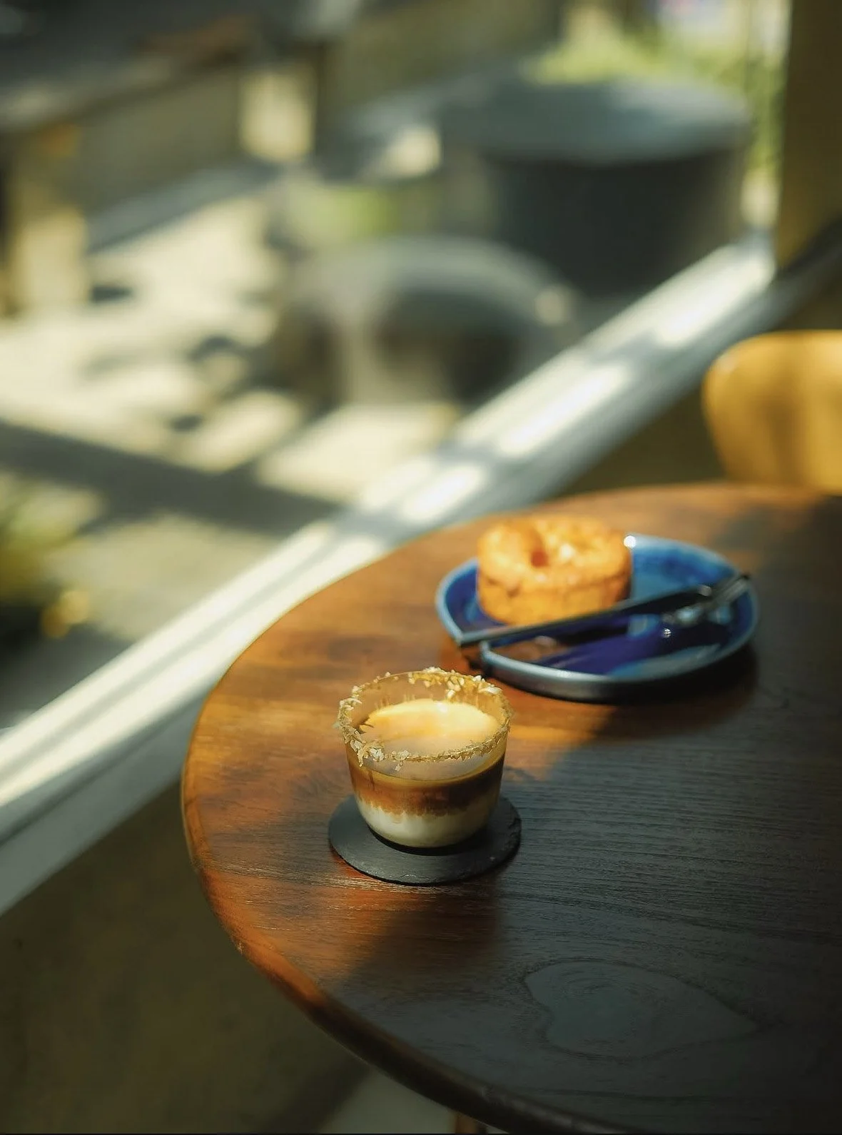 A dessert in a glass with layers of cream, caramel, and chocolate, garnished with a caramelized sugar rim, placed on a dark coaster on a wooden table. In the background, there is a plate with a donut and a slice of cake, and a blue plate with a fork 