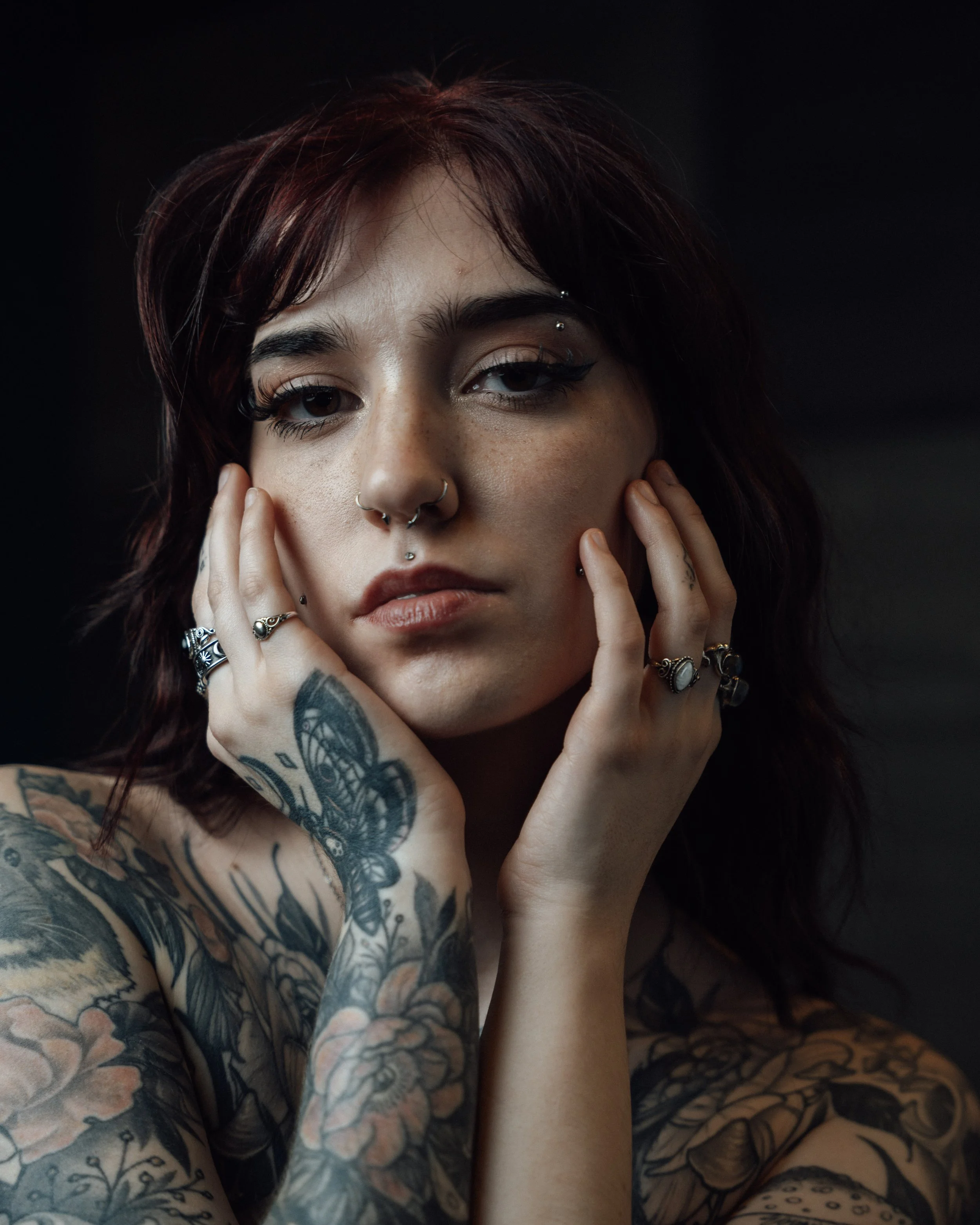 Close-up portrait of a young woman with short dark red hair, multiple facial piercings, tattoos on her arm and shoulder, wearing rings and makeup, against a dark background.