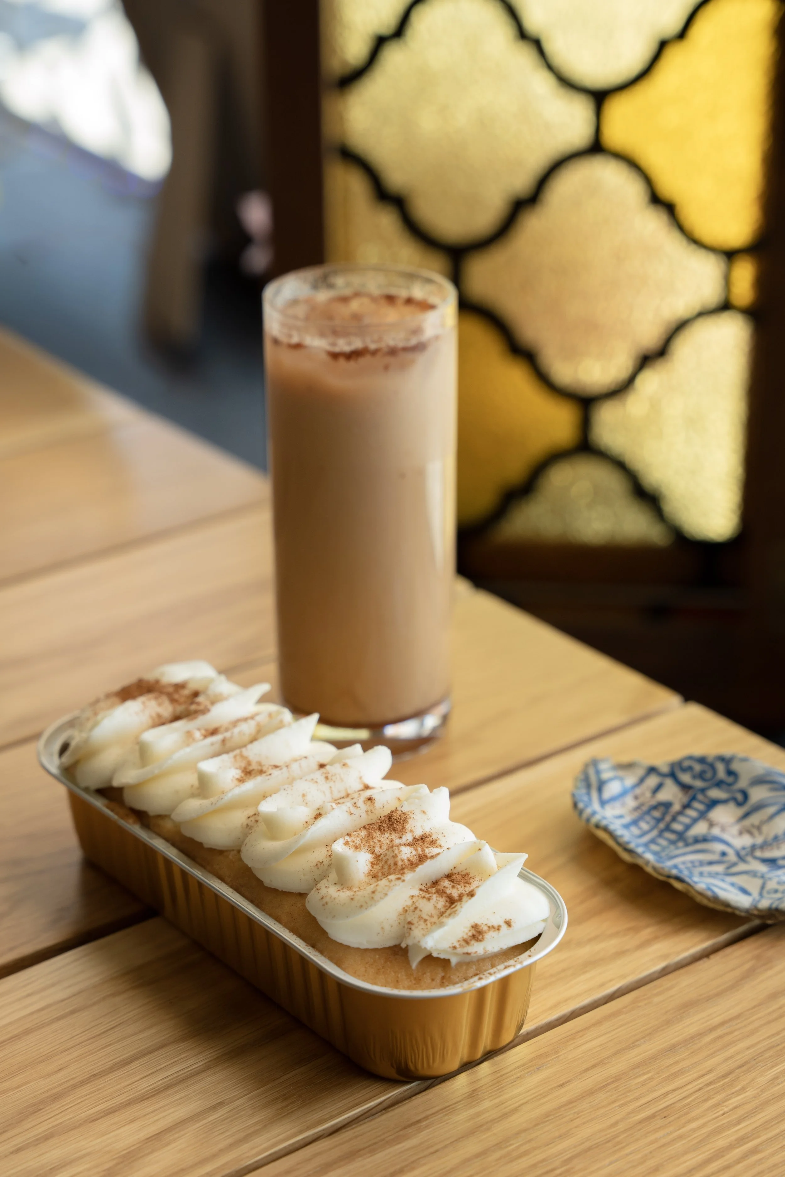 A dessert with whipped cream topped with cinnamon in a rectangular container, and a tall glass of iced coffee with milk and cocoa powder, on a wooden table with a decorative window in the background.