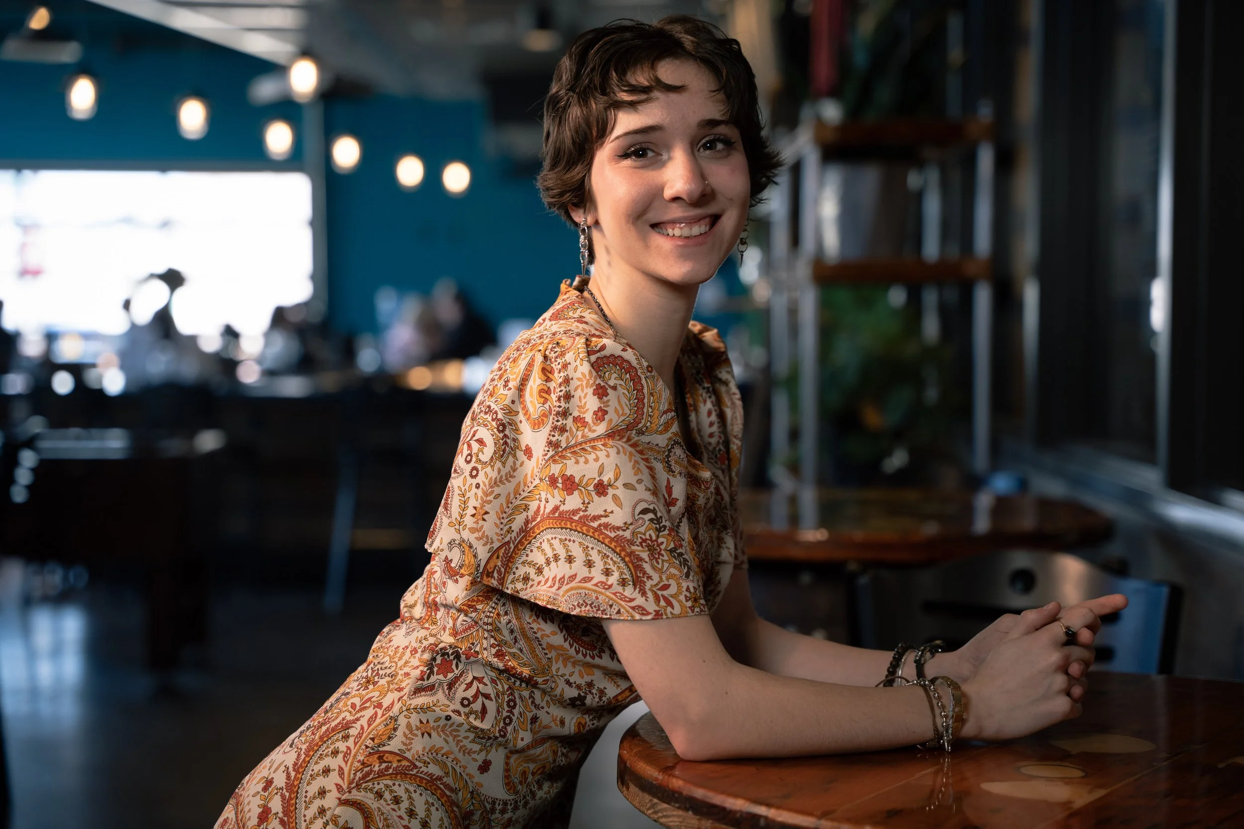 A young woman with short brown hair and earrings smiling while sitting at a wooden table in a cozy, dimly-lit cafe with blurred background.