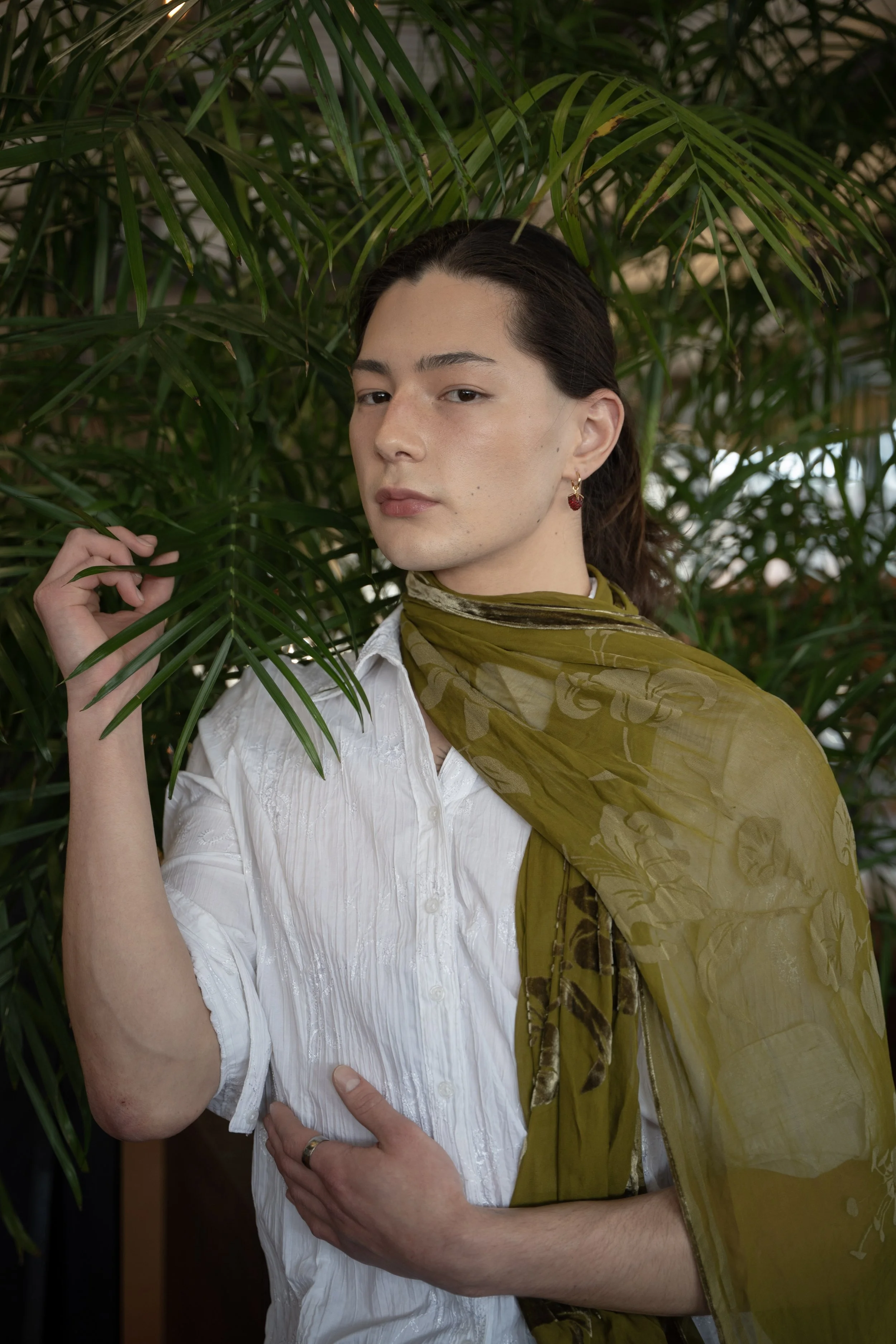 A person with dark hair, wearing a white shirt and an olive green scarf, standing among green palm leaves.