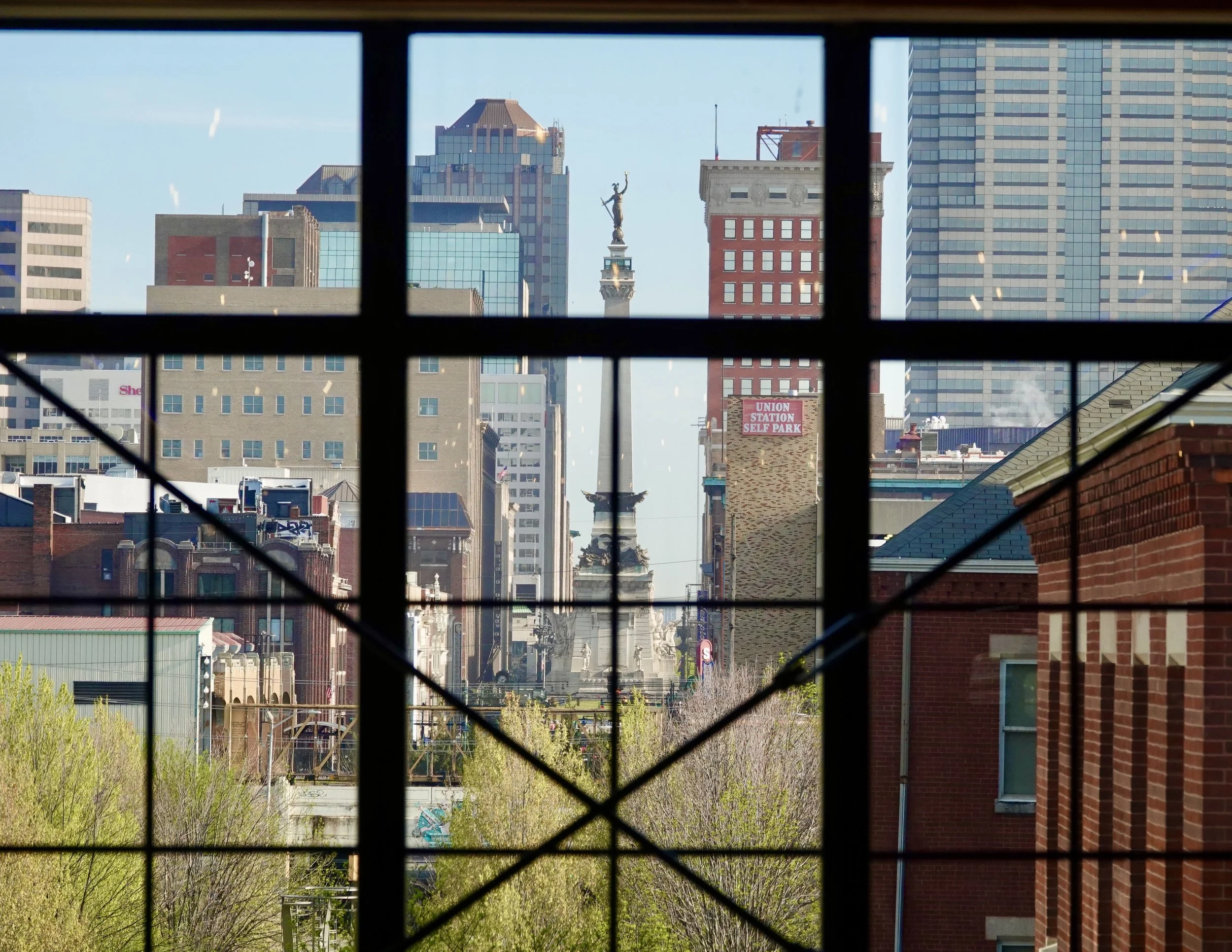 City skyline viewed through a window with black grid bars, featuring tall modern skyscrapers and a monument with a statue in the center.