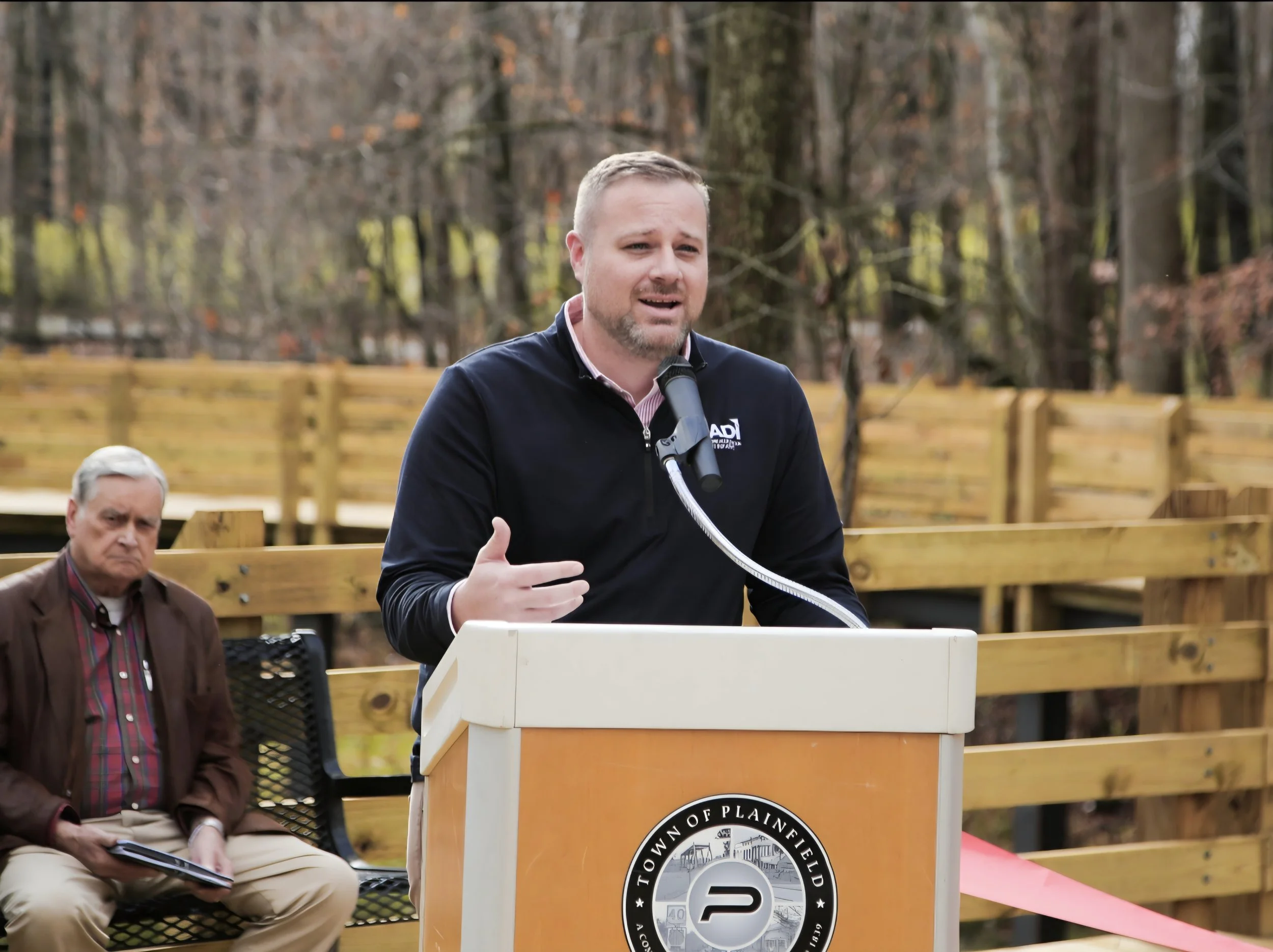 A man speaking at a podium outdoors, with two other men seated nearby. The background features trees and a wooden fence, indicating a park or public space during fall.