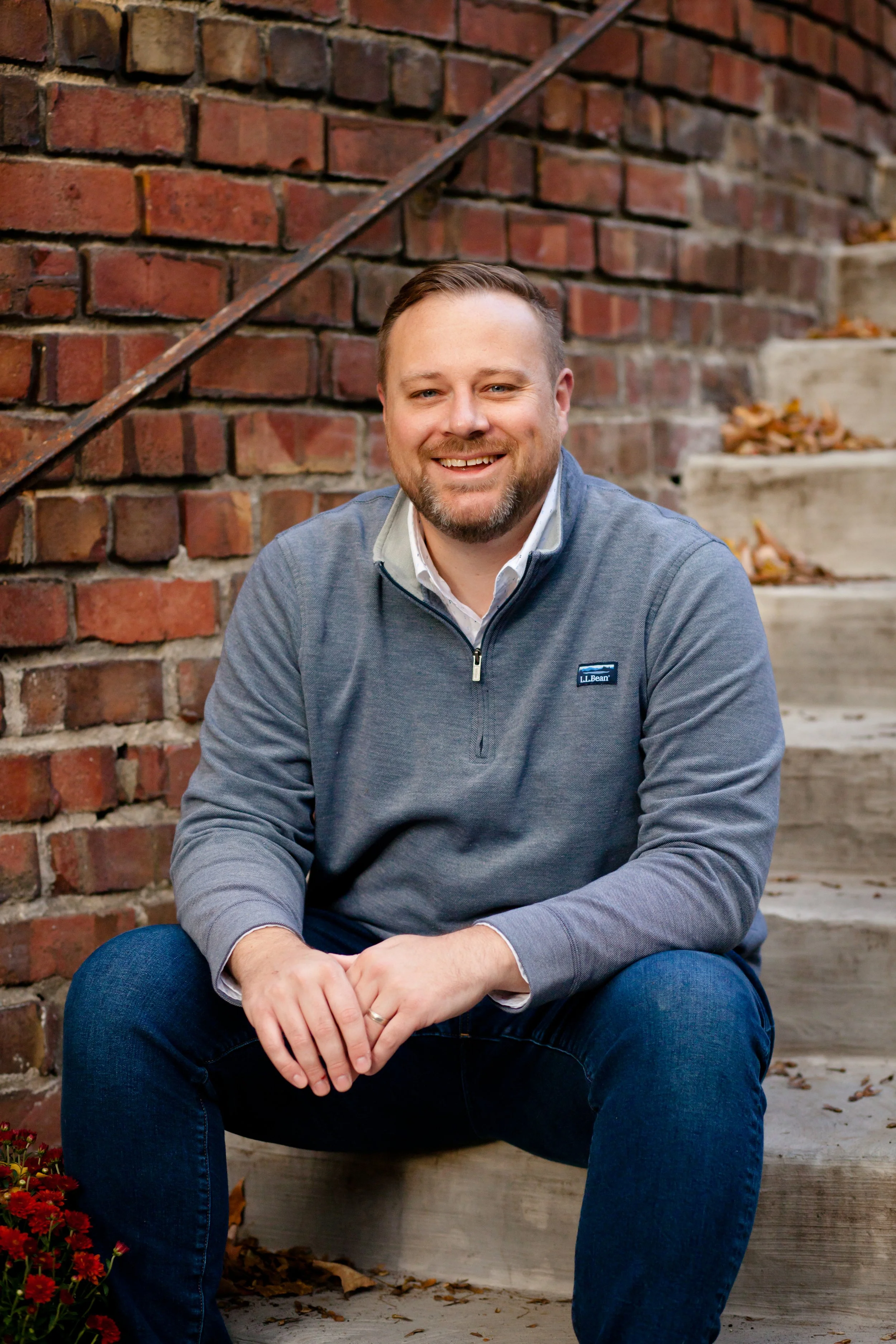 A smiling man with a beard, wearing a gray pullover and blue jeans, sitting on steps beside a brick wall, with fallen leaves and flowers around.