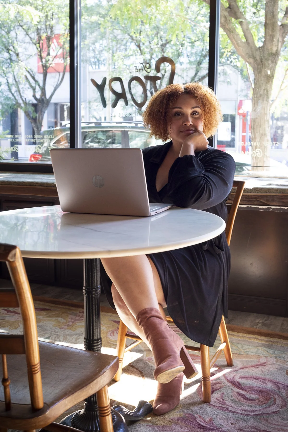 A woman with curly hair sitting at a round table inside a cafe, looking at the camera with her chin resting on her hand, with a laptop in front of her and large windows behind displaying trees and the street.