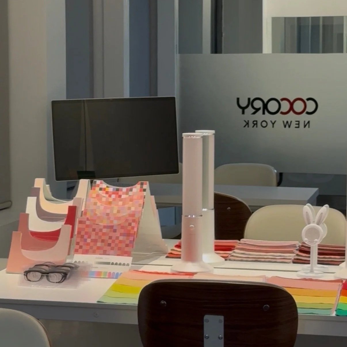 A desk with color samples, a computer monitor, a white lamp, a pair of black glasses, and rainbow-colored paper strips. In the background, a glass wall with the logo 'COCORY NEW YORK' is visible.