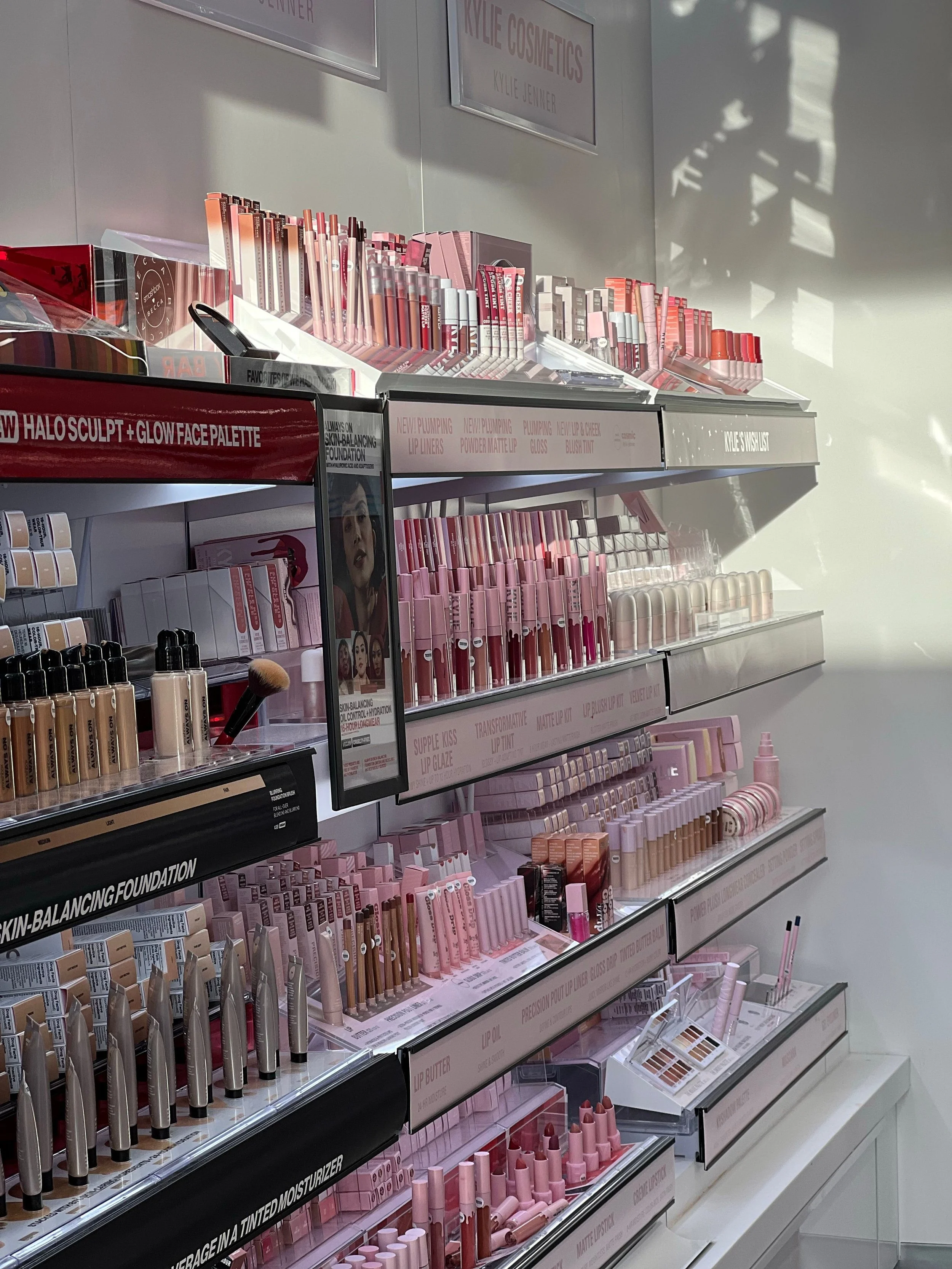 Display shelves with pink and neutral-colored makeup products such as lipsticks, lip glosses, and foundations in a cosmetics store.
