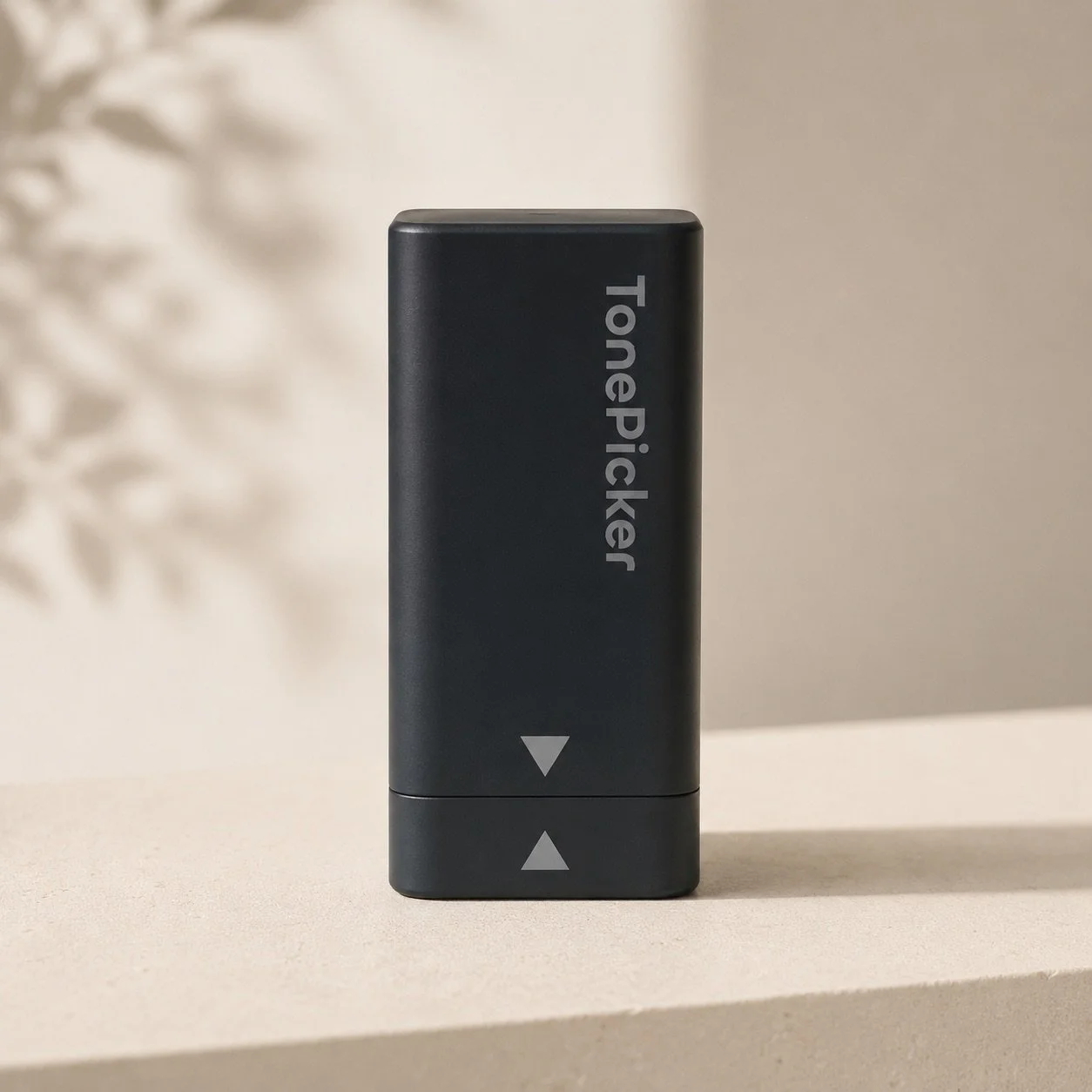 A black TielPickler device with arrow buttons on a beige surface, with a blurred decorative plant in the background.