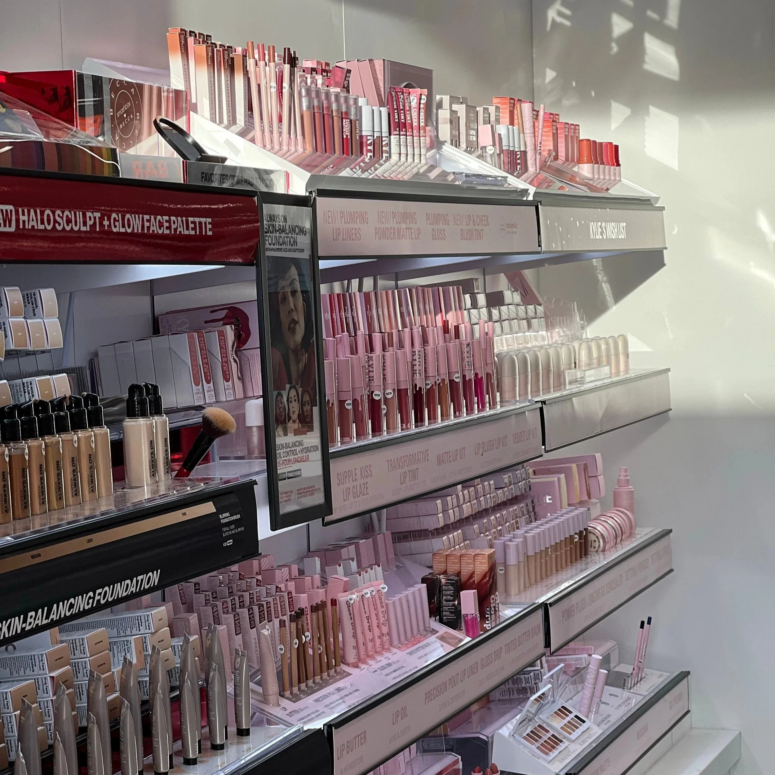 Beauty products such as lip gloss, lip liners, foundation, and face palettes arranged on store shelves with pink and neutral packaging.