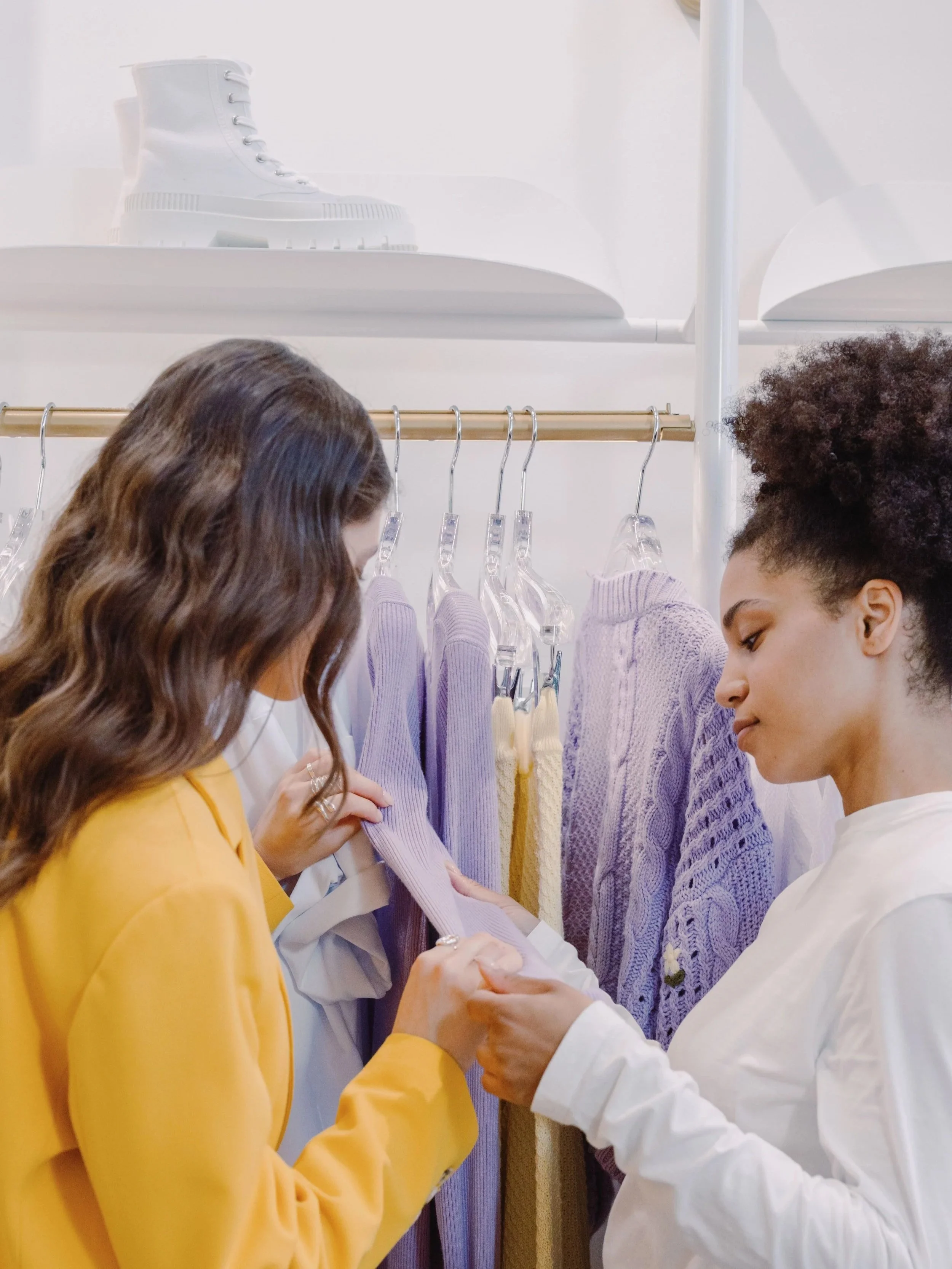 Two women shopping for sweaters at a clothing store, holding a purple sweater between them, with sweaters hanging on a rack behind them.